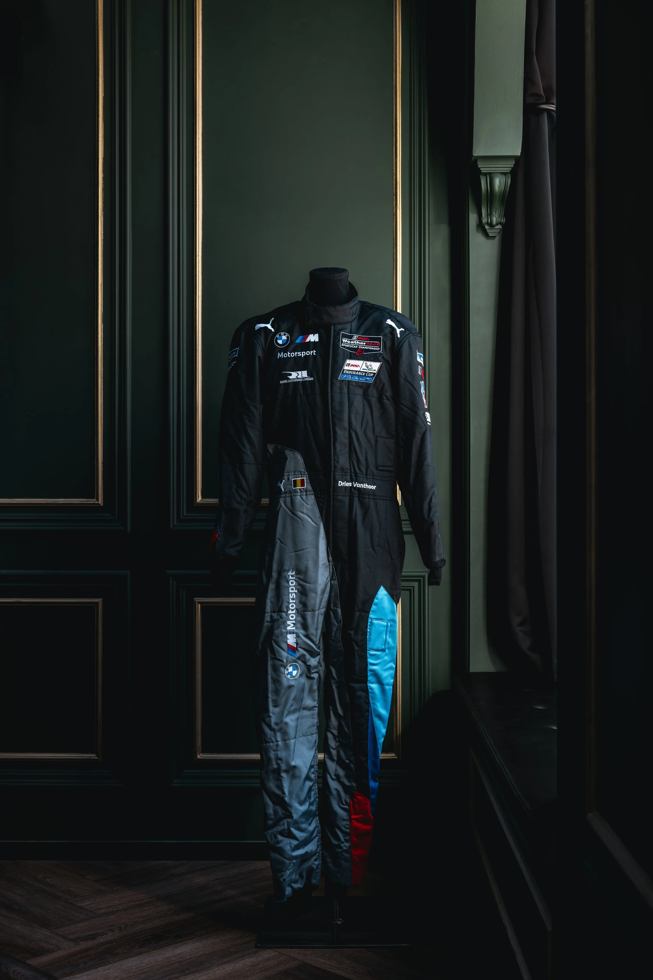 A racing driver’s suit displayed on a mannequin, with sponsorship logos including BMW, Puma, and others, set against a dark green paneled wall with gold accents.