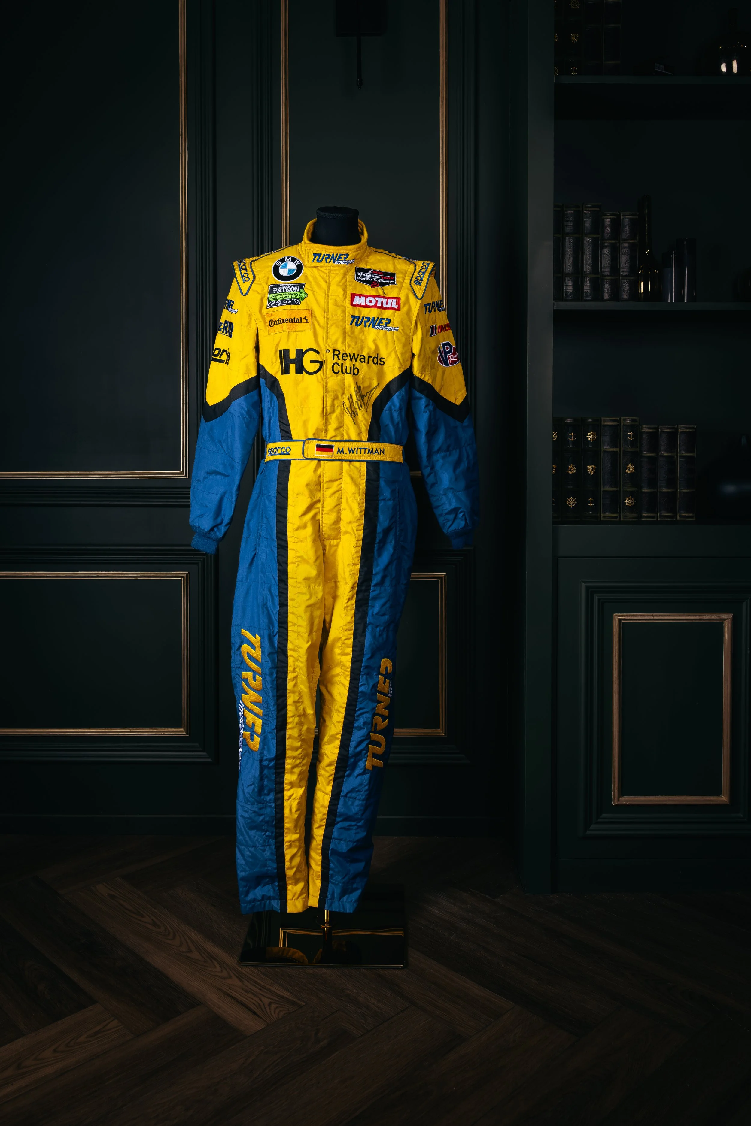 A racing suit with yellow and blue colors displayed on a mannequin, featuring various sponsor patches and the name M. Wittmann.