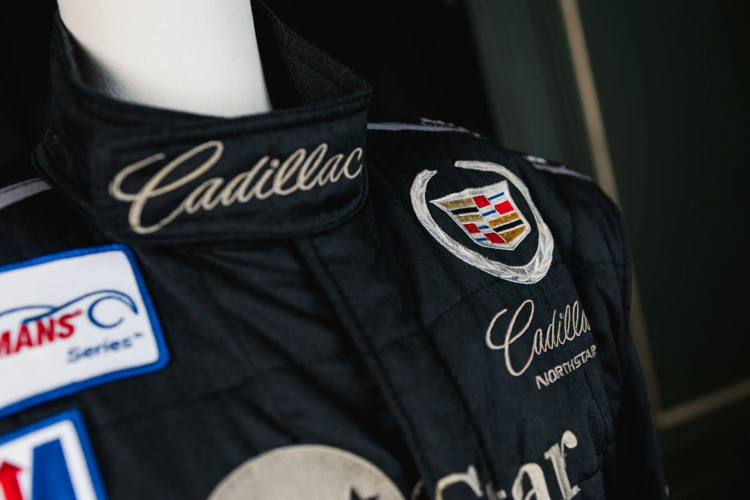 Close-up of a black racing jacket with Cadillac logo and patches, including 'Cadillac' embroidery and a 'Lexus Motors' patch.