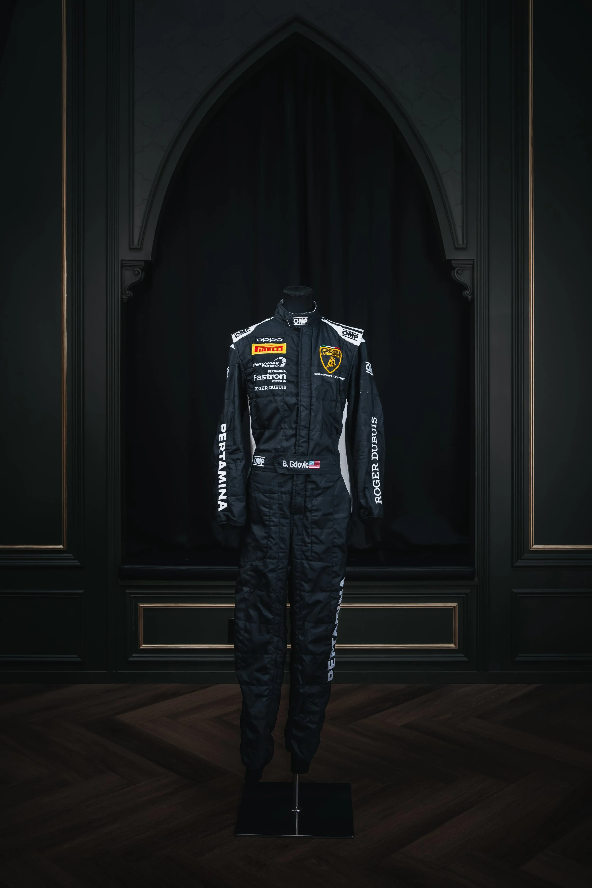 A black racing suit on a mannequin with Lamborghini and sponsor logos, displayed in a dark, elegant room.