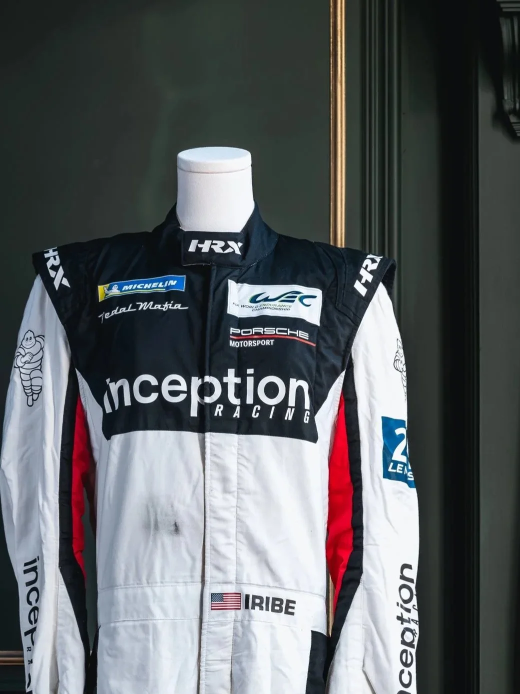 Racing suit displayed on a mannequin with various sponsor logos, including Michelin, Porsche Motorsport, and Inception Racing, and an American flag patch labeled 'Iribe'.