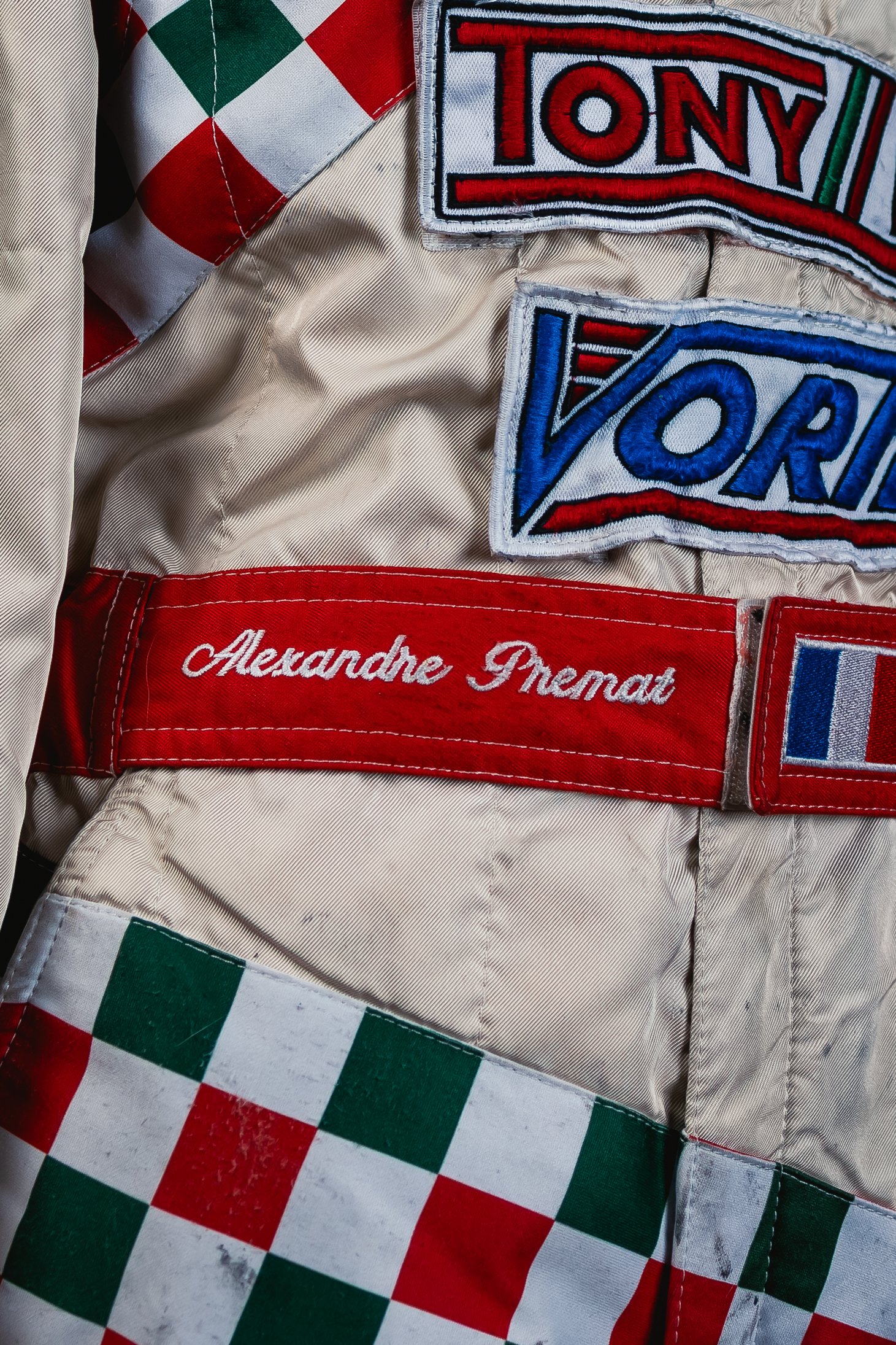 Close-up of a racing jacket featuring patches with the names 'Tony', 'Vord', and 'Alexandre Premat', along with a red, white, and green checkered pattern.