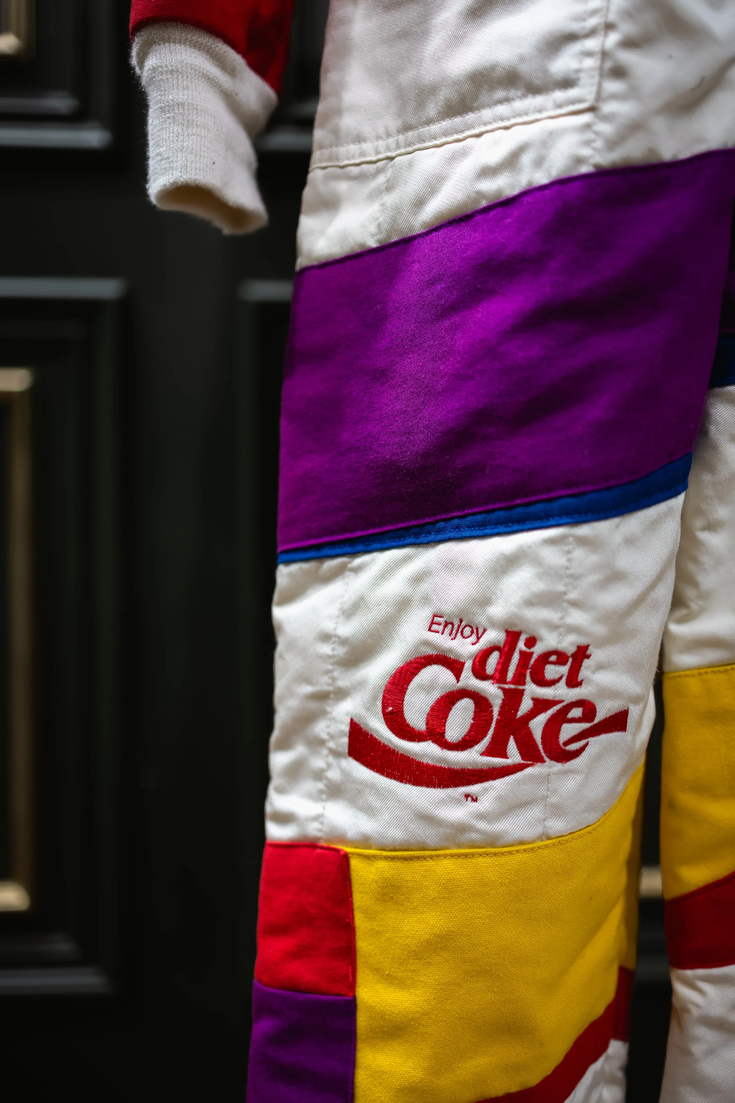 Close-up of pants with multiple colored fabric patches, including a whitepatch with a red "Diet Coke" logo and the phrase "Enjoy diet Coke".
