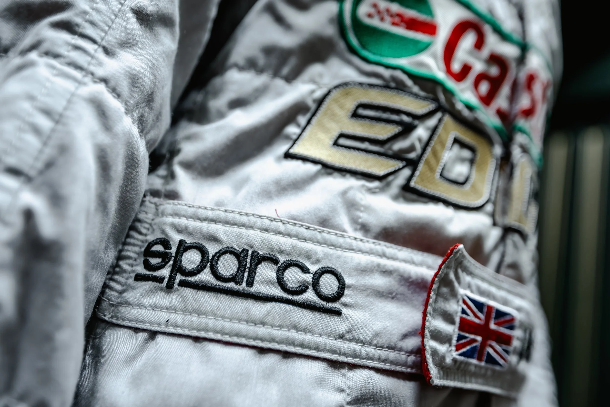 Close-up of a racing suit with patches including the name 'Sparco,' a Union Jack flag, and logo patches.