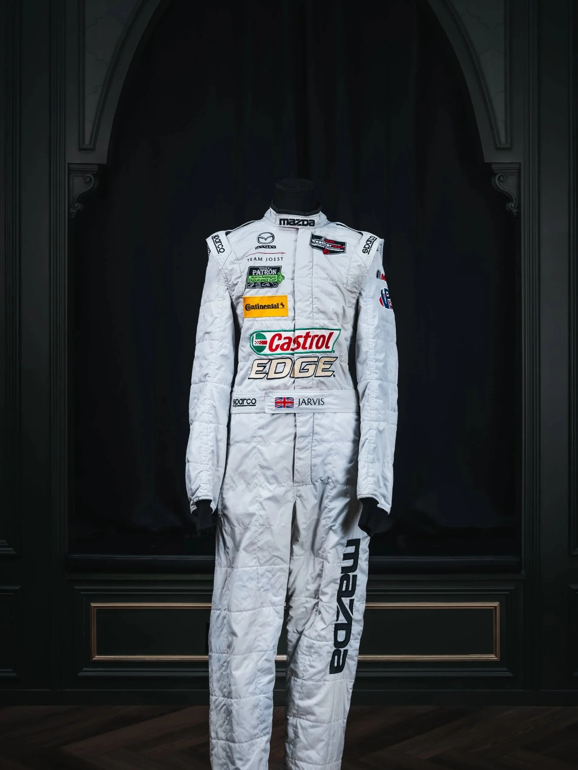 A white racing suit with various sponsor logos displayed on a mannequin, standing against a dark background.