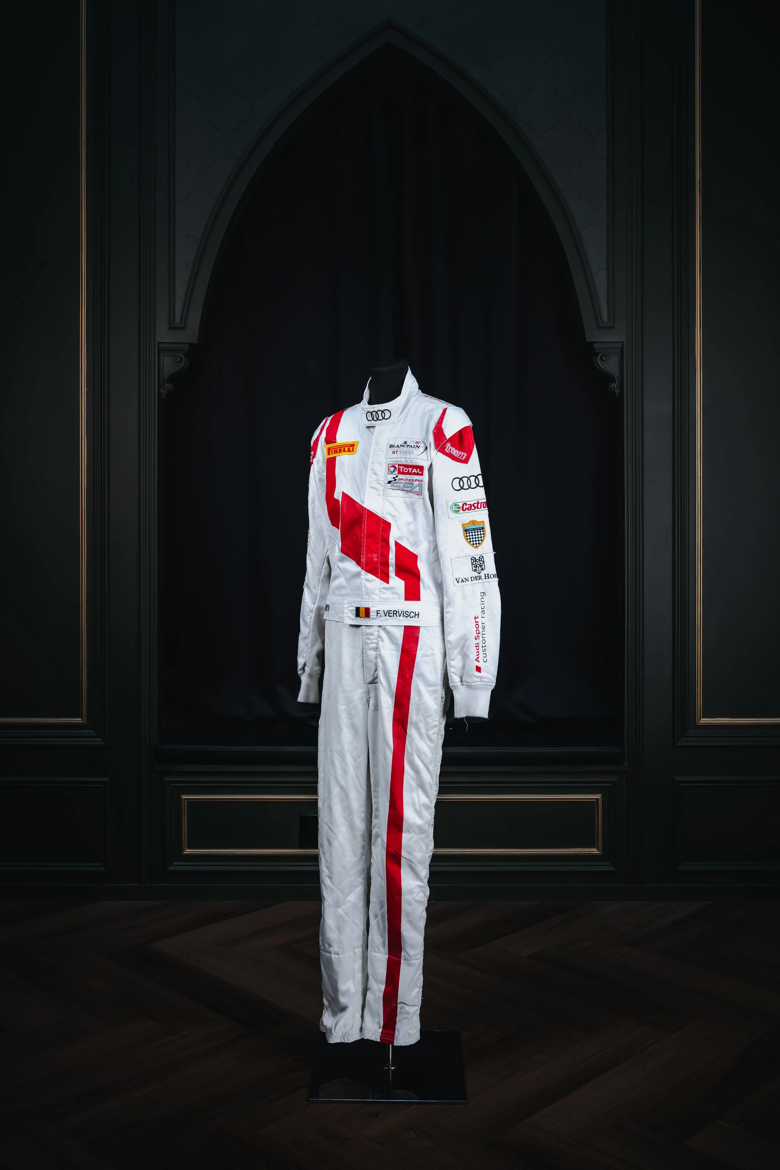 A racing suit on display in a dark exhibit with black walls and wooden flooring.