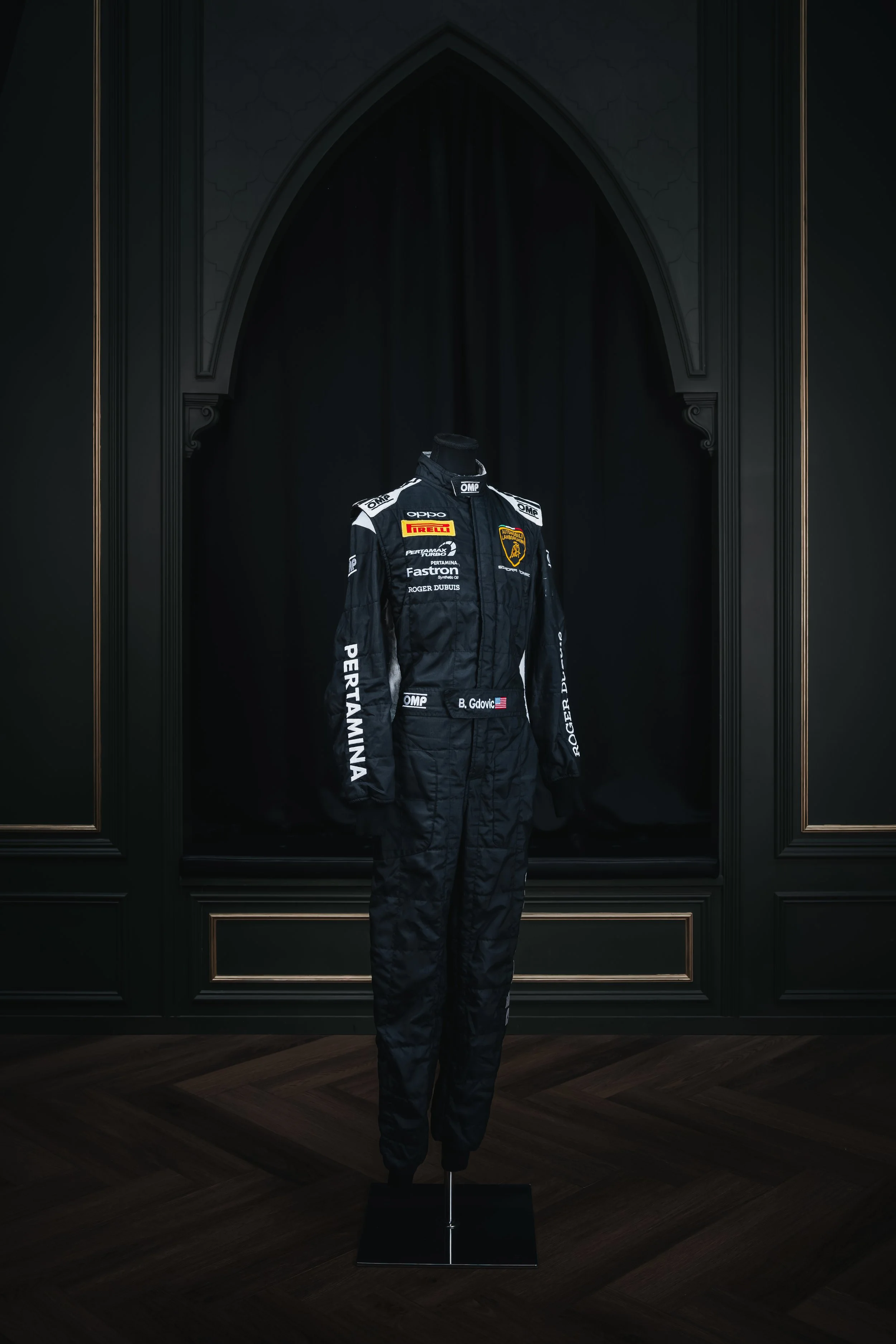 A black racing suit with various sponsors, including Lamborghini and Pirelli, displayed on a mannequin against a black backdrop in an elegant room with dark wood floors and black paneled walls.