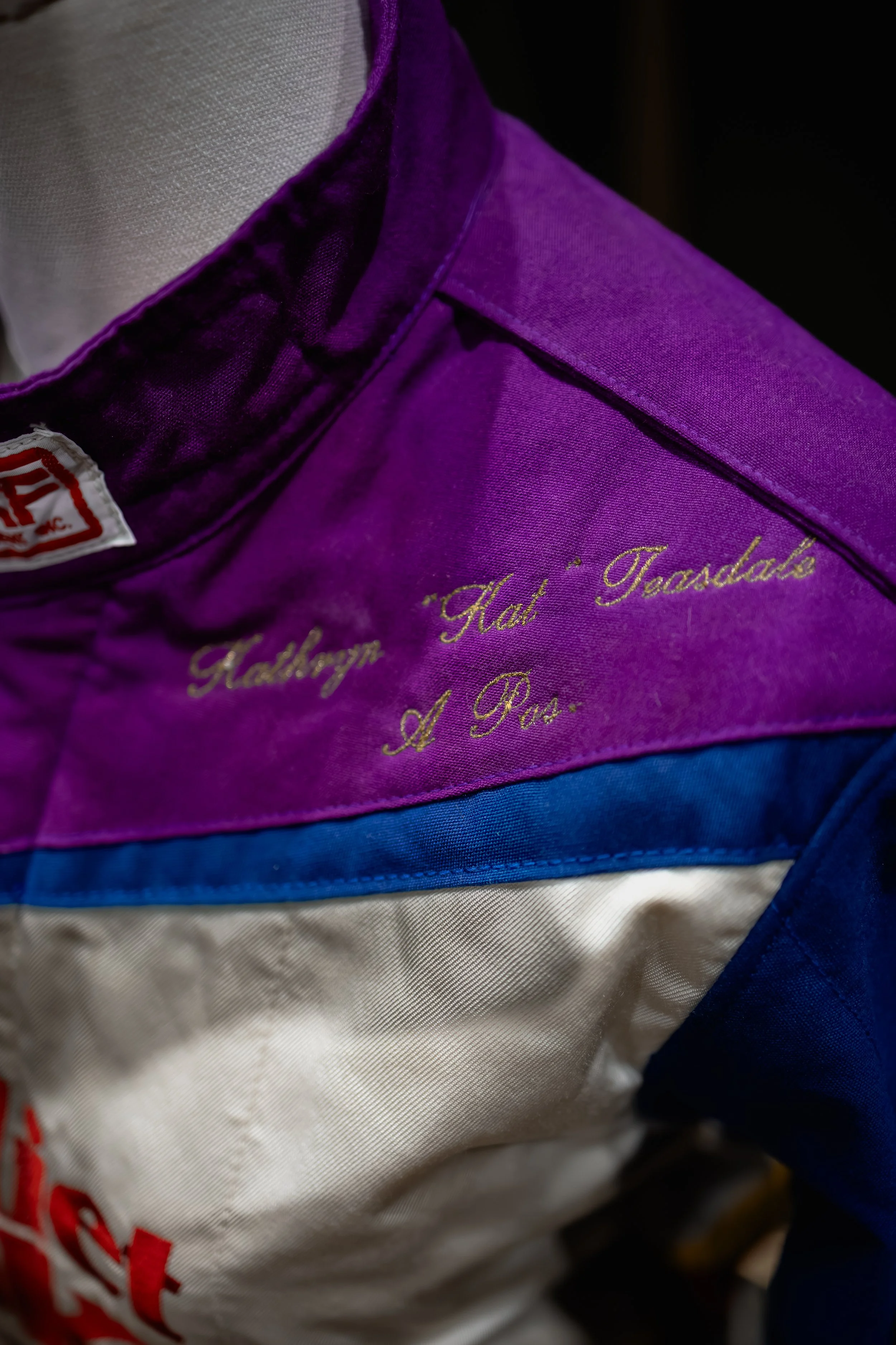 Close-up of a colorful jacket showing embroidery that reads 'Hab. 'Tadalah' A Pas.' on a purple section, with parts of a tag and other fabric sections in white, blue, and red.