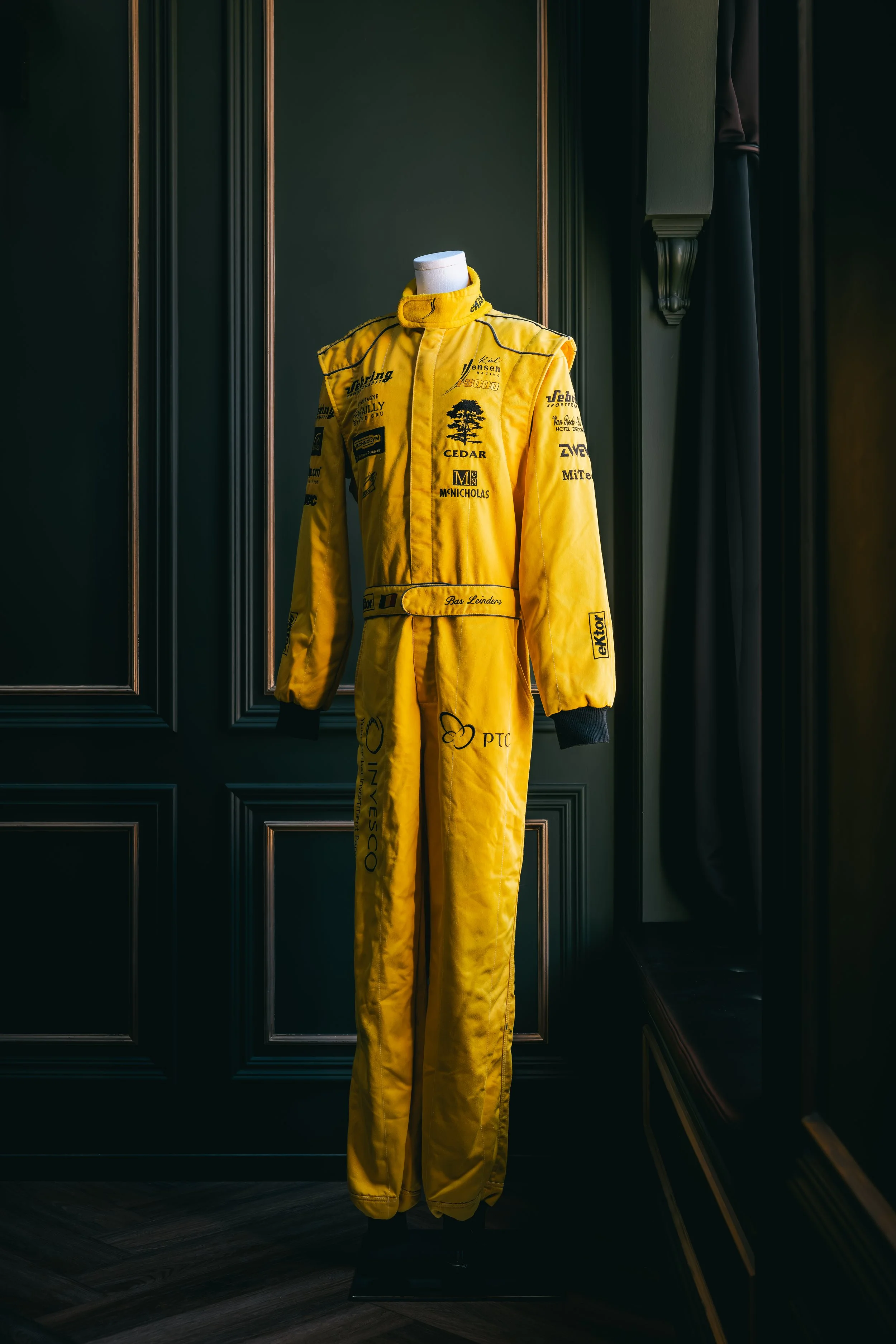 Yellow racing suit with various sponsor logos displayed on a mannequin in a dark room.