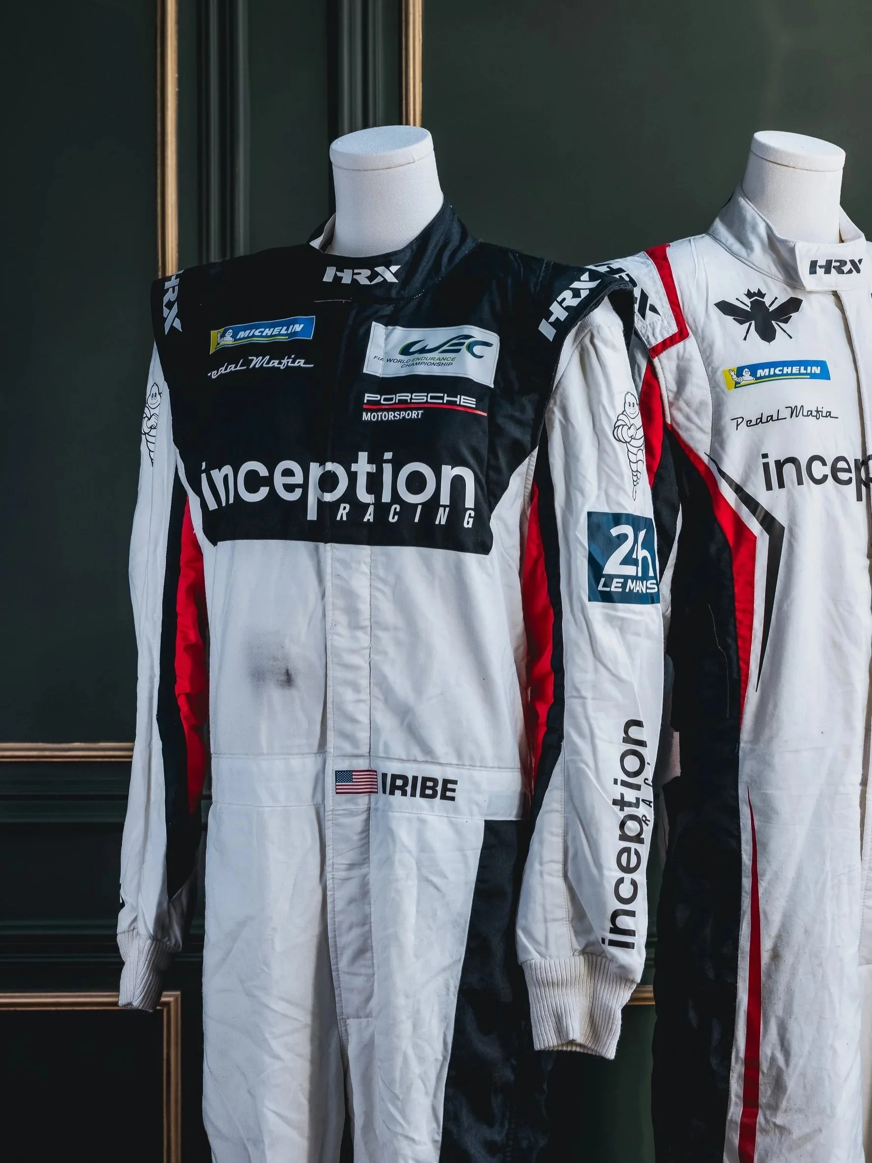 Two racing suits on mannequins with various sponsor logos, one black and white with red accents, the other white with black and red accents.