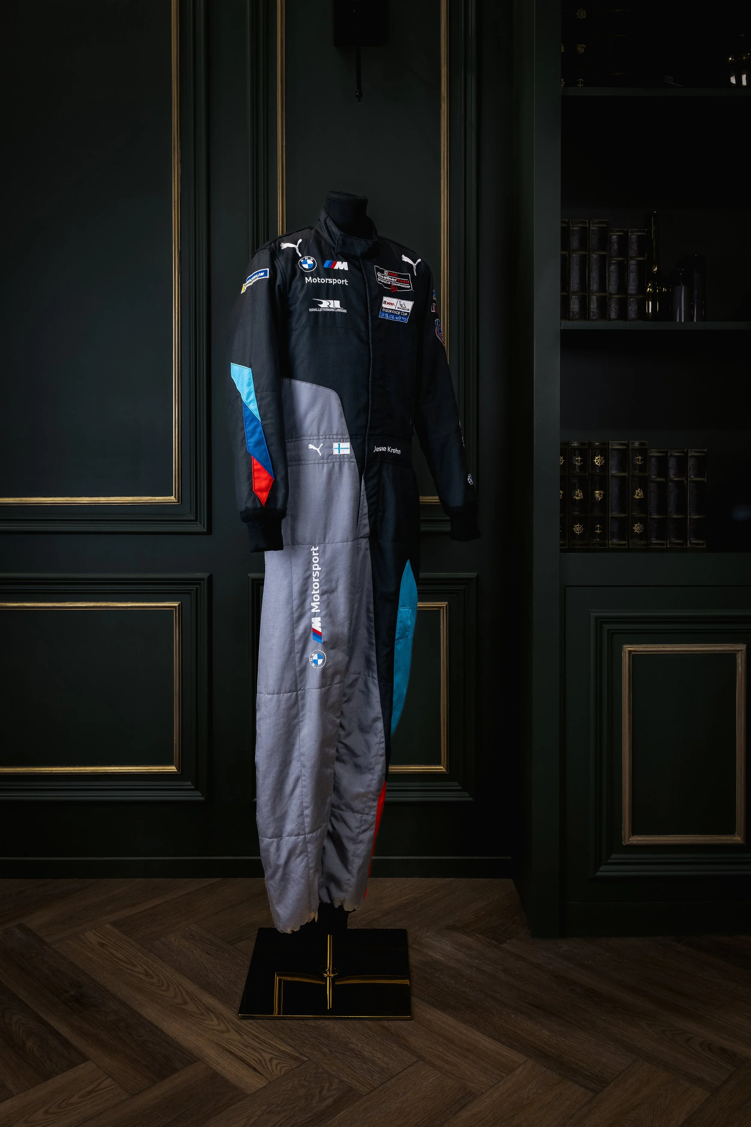 A racing suit displayed on a mannequin in a room with dark green walls and wooden flooring.