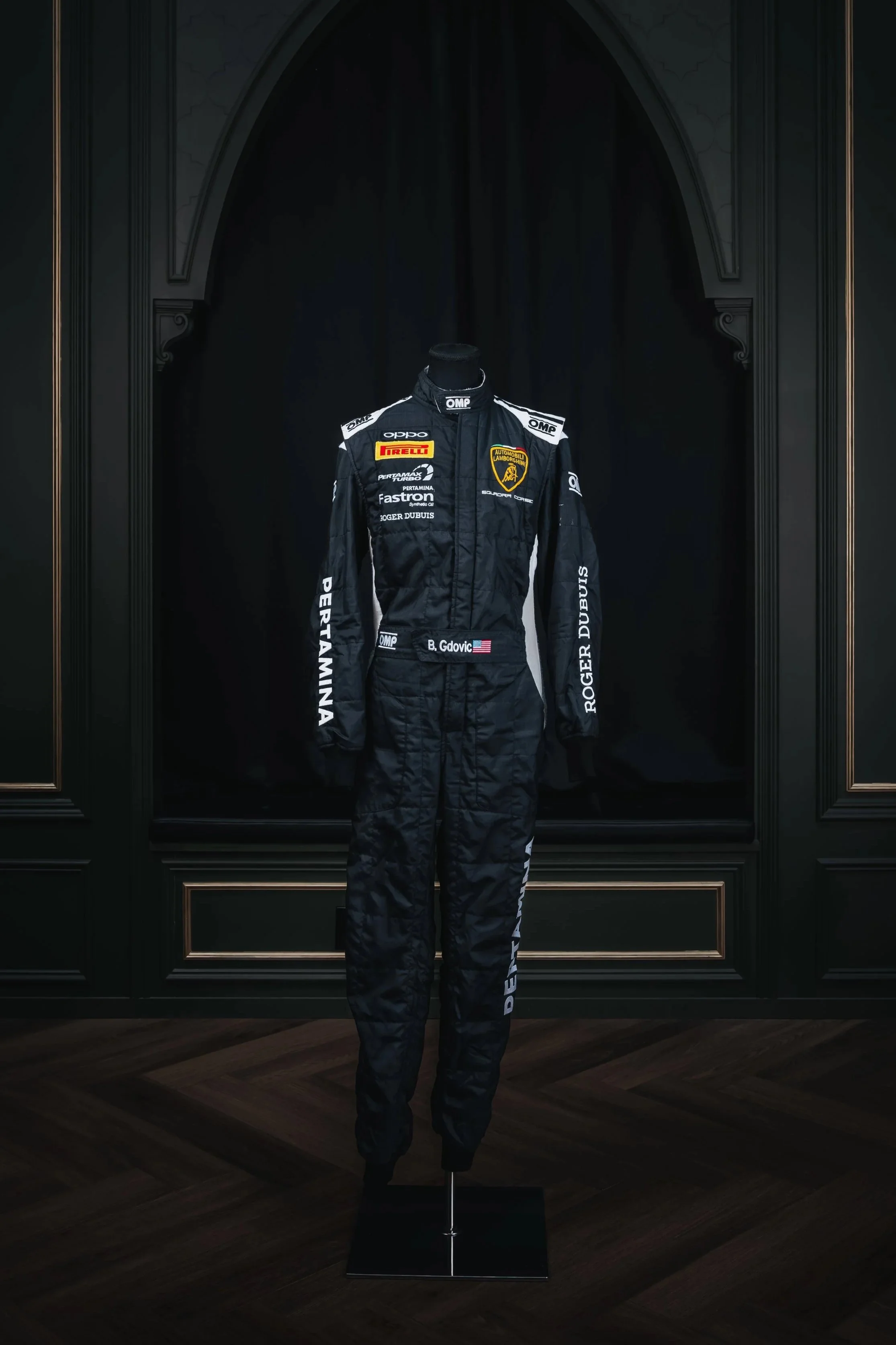 A black racing suit with sponsor patches and the Lamborghini logo displayed on a mannequin in front of a dark backdrop.