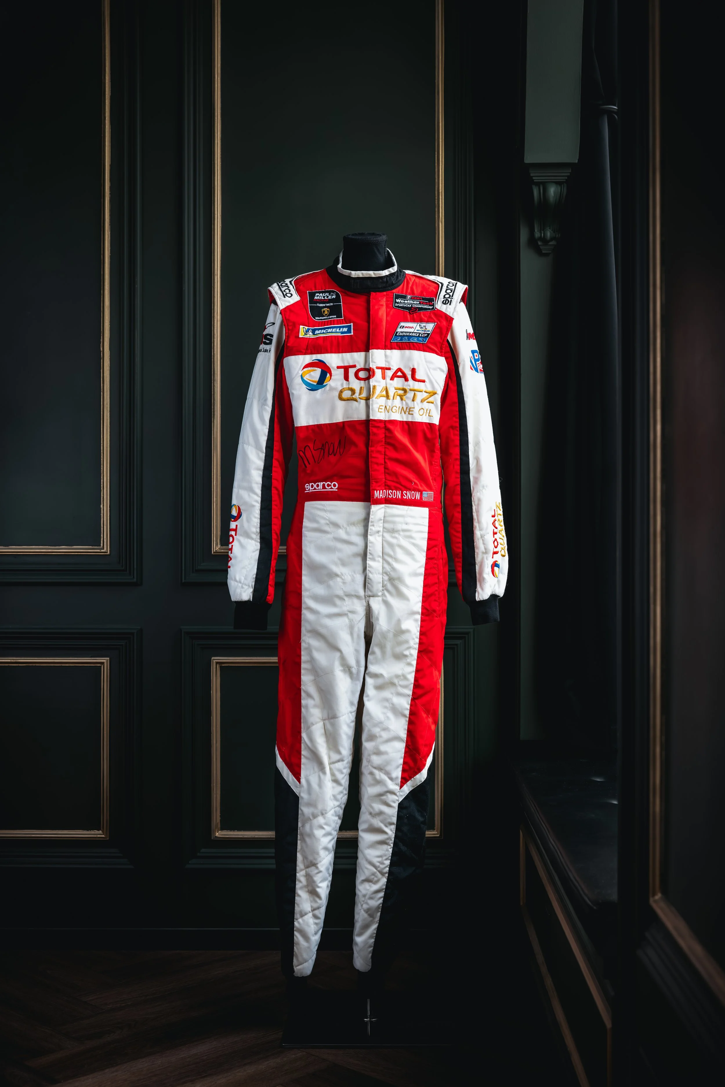 Red and white racing suit displayed on a mannequin, with various sponsor patches, including Total Quartz Engine Oil, in a dark room with ornate black and gold wall paneling.