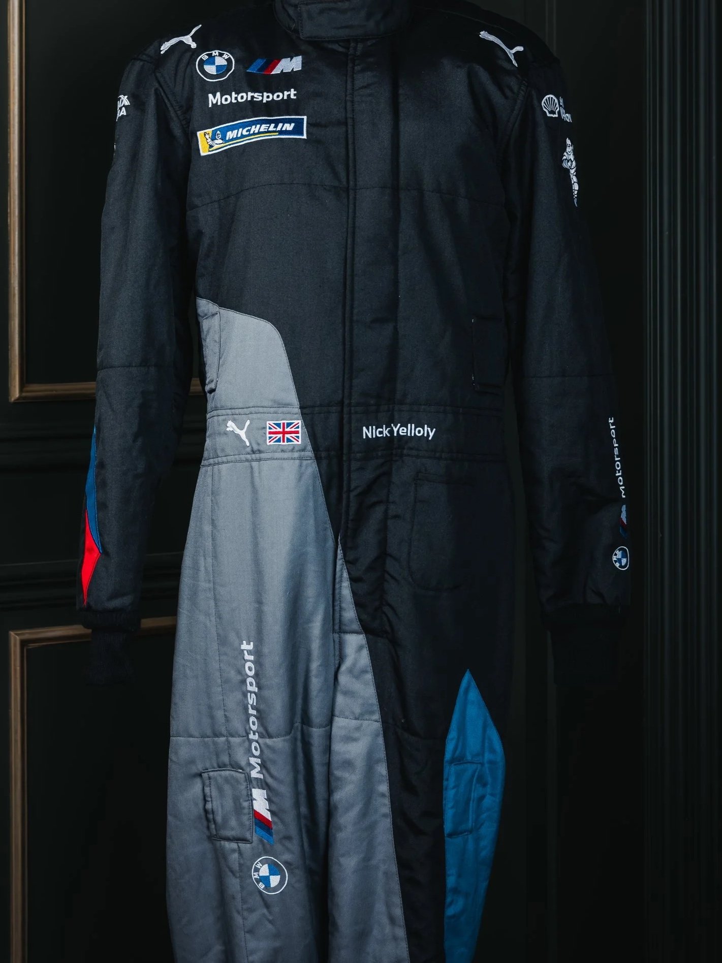 Dark racing suit with multiple sponsor logos, including BMW, Michelin, Puma, and Shell, displayed on a mannequin.