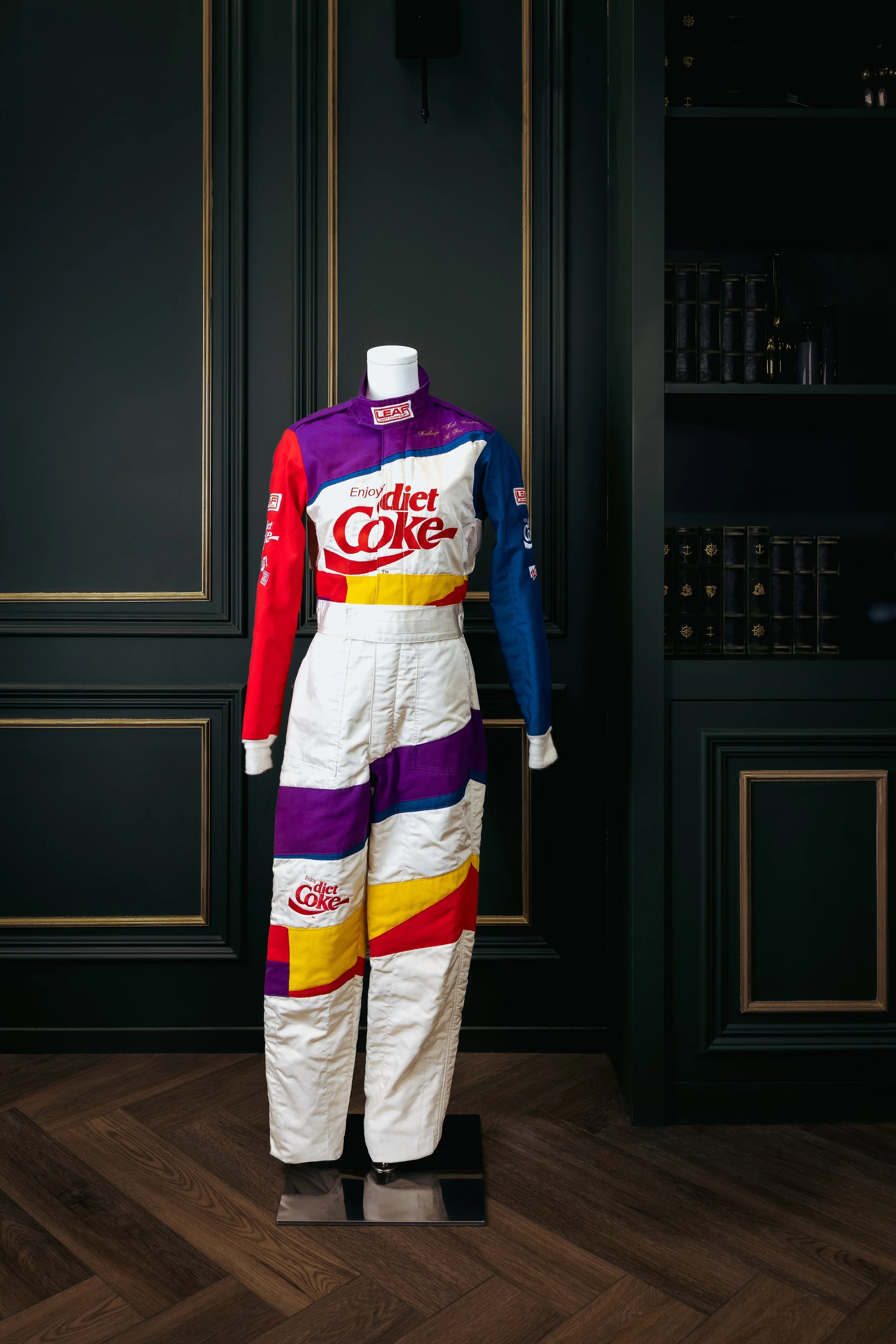 Colorful vintage race suit with sponsors and logos, displayed on a mannequin in a dark room with wooden floors and dark green paneled walls.