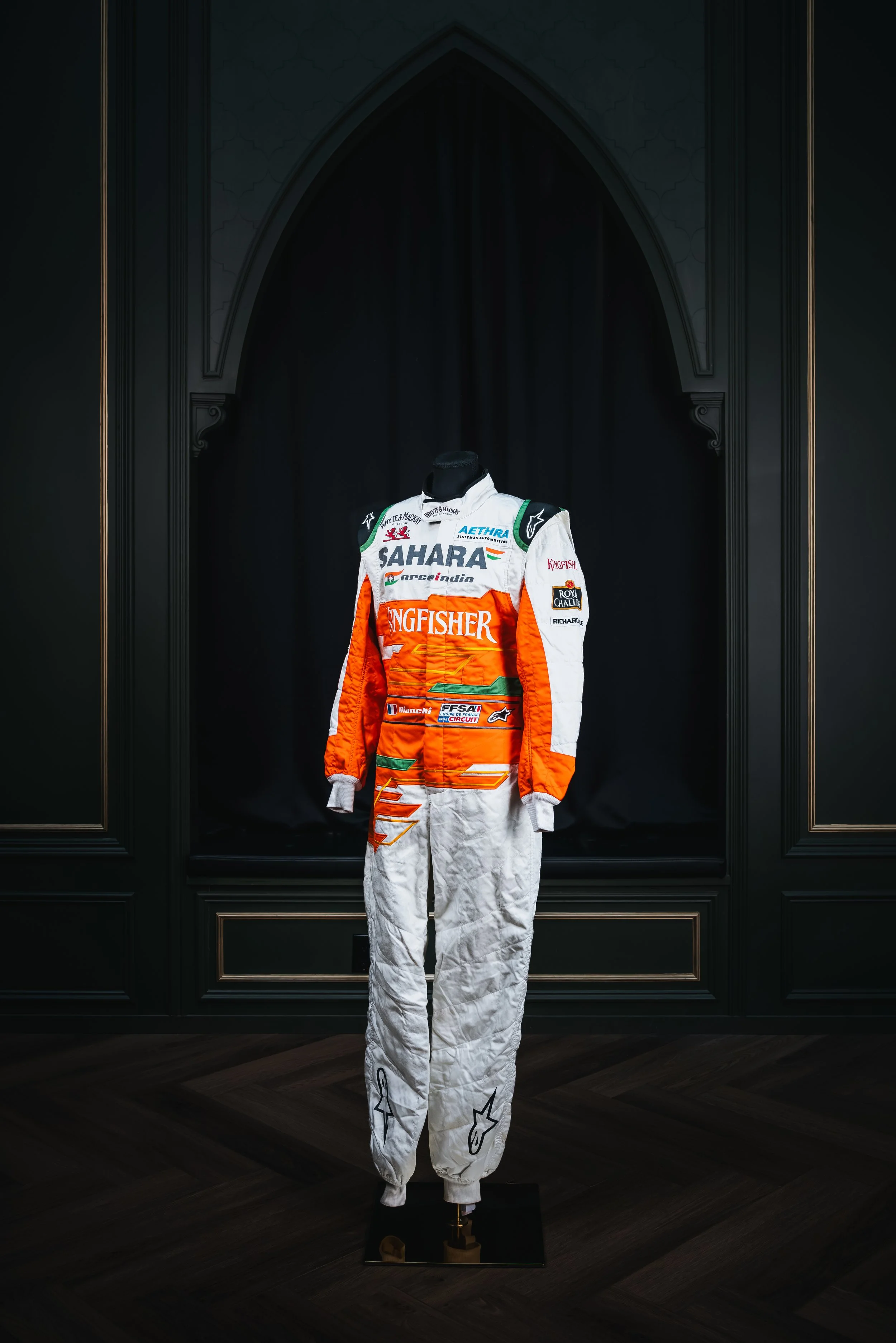 Motorsport racing suit on display against black background with ornate black walls and wooden floor.
