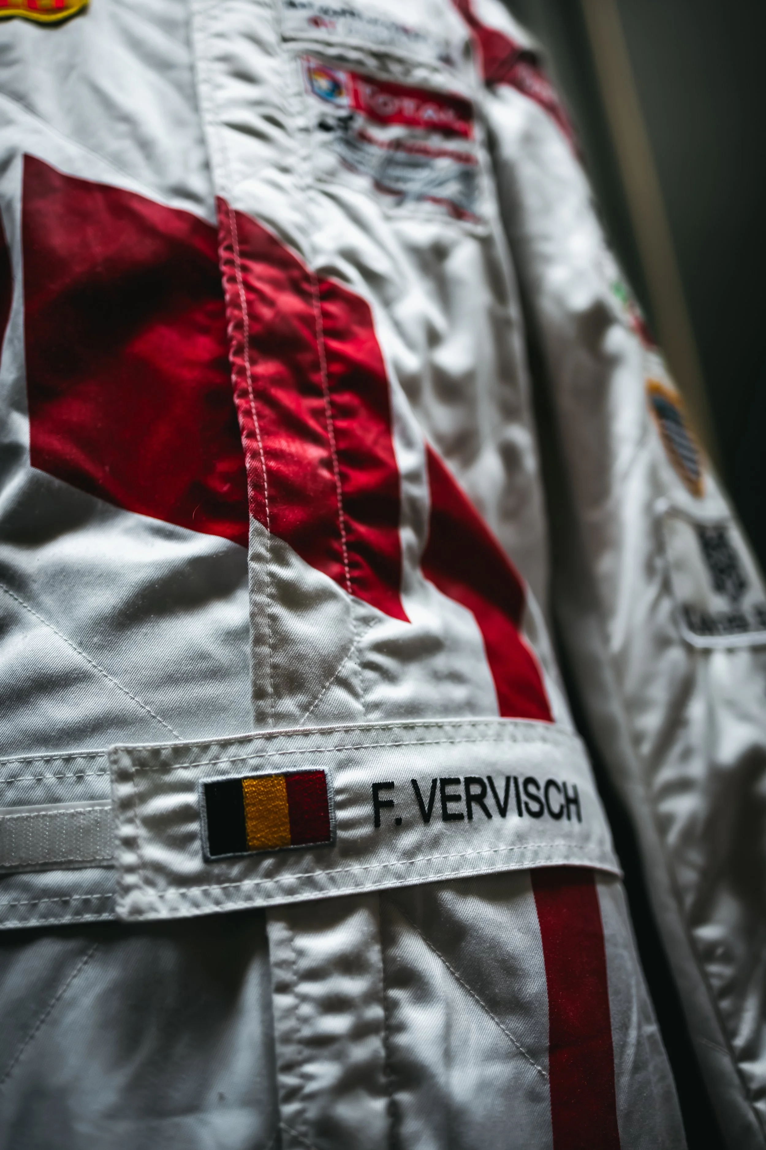 Close-up of a racing suit with a Belgian flag, the name F. VERVISCH, and various sponsor patches, likely belonging to a race car driver.