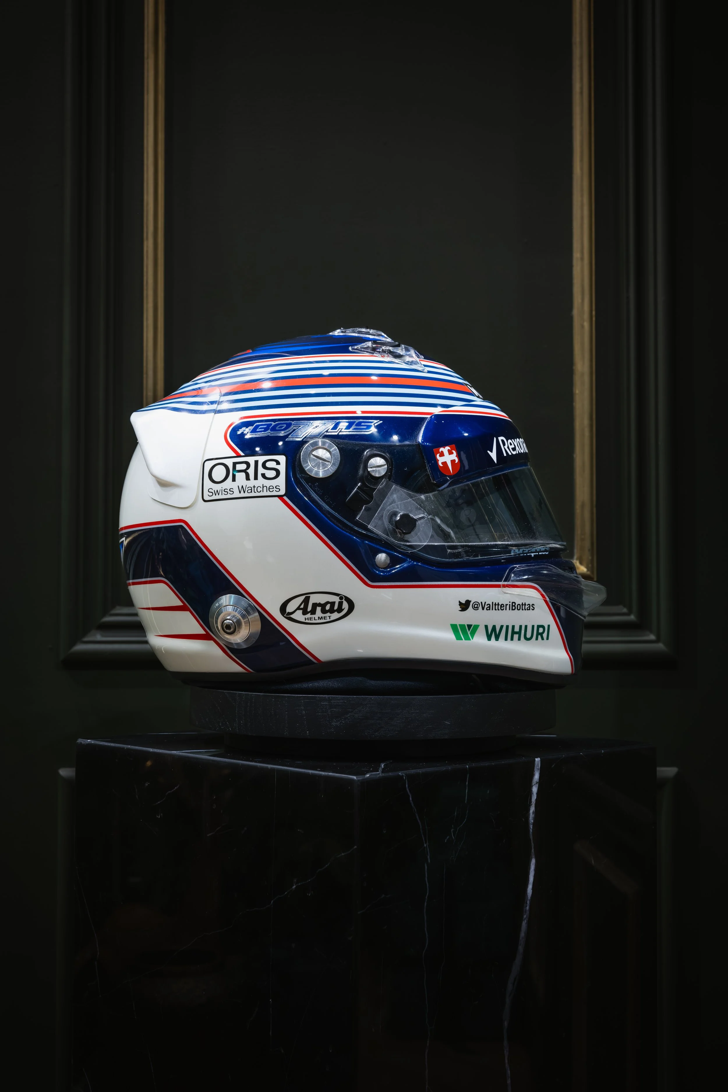 A racing helmet with a blue, white, and red design on a black pedestal, set against a dark background.