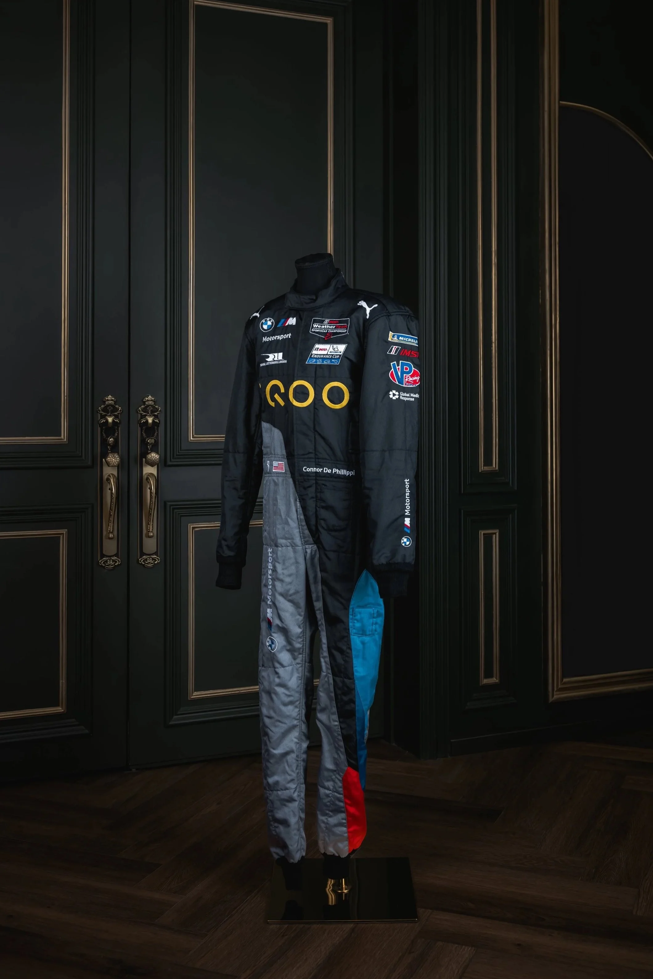 A smiling race car driver in a black racing suit with sponsor logos, in a garage or pit area, with a helmeted figure and garage environment in the background.