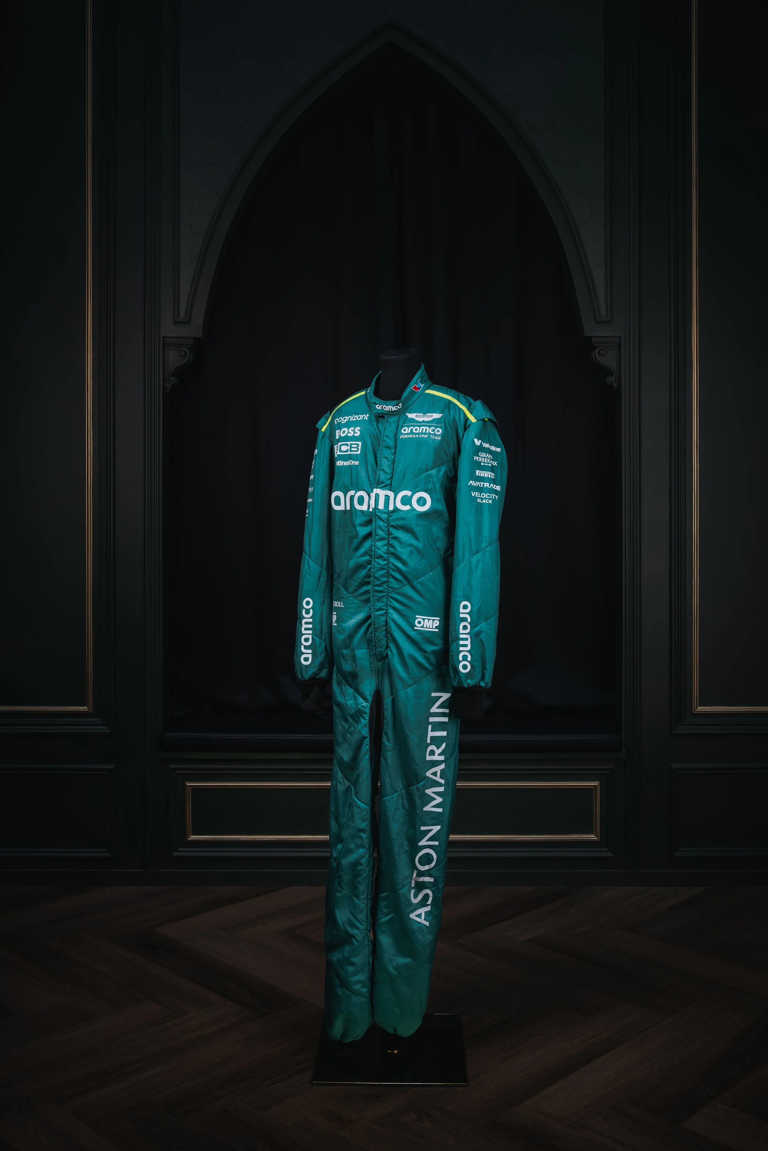 A teal racing suit with sponsors' logos, displayed on a black mannequin, with 'ASTON MARTIN' written on the left leg.