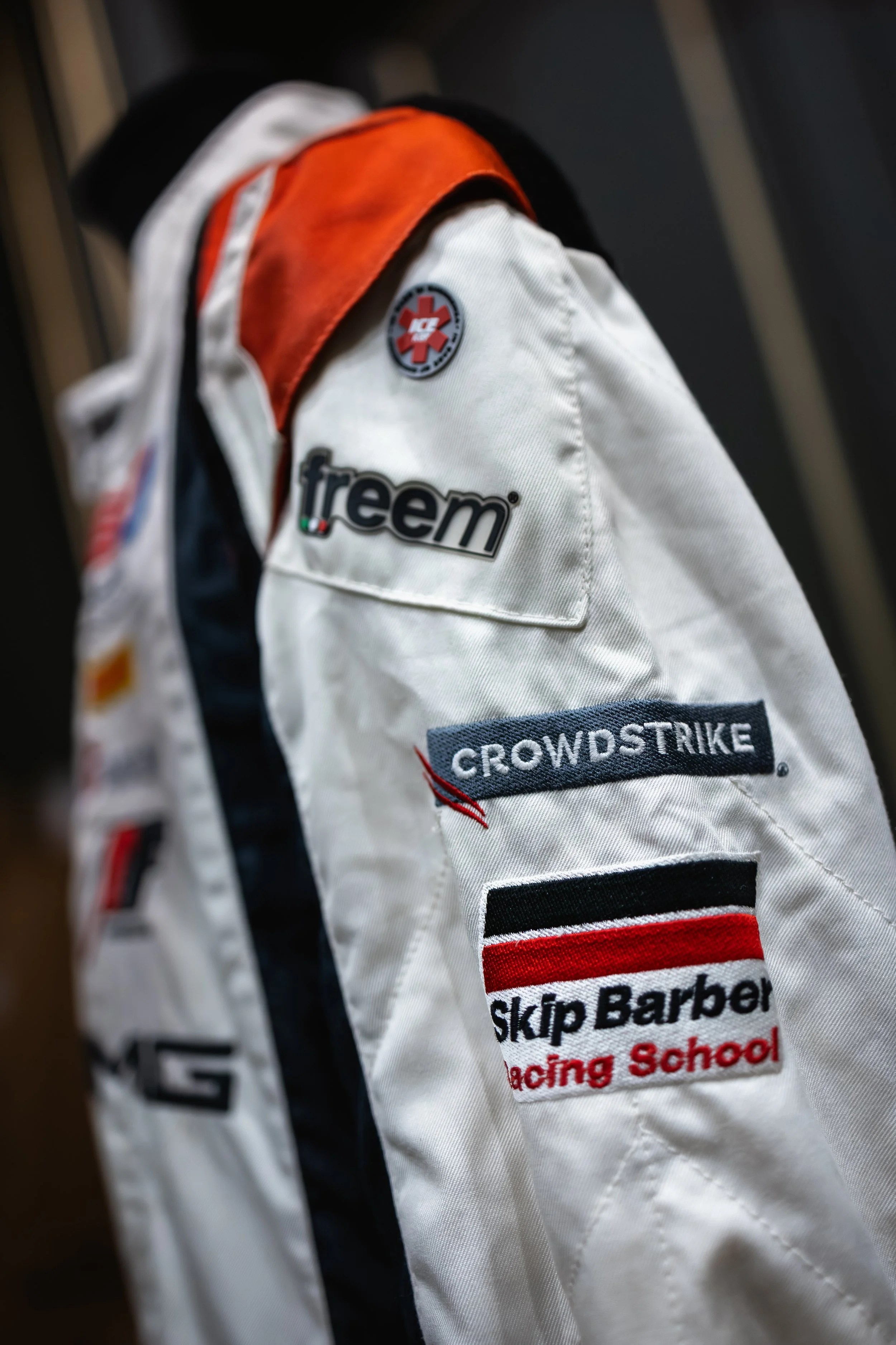 Close-up of a racing jacket with multiple sponsor patches including 'freem,' 'CrowdStrike,' and 'Skip Barber Racing School.'