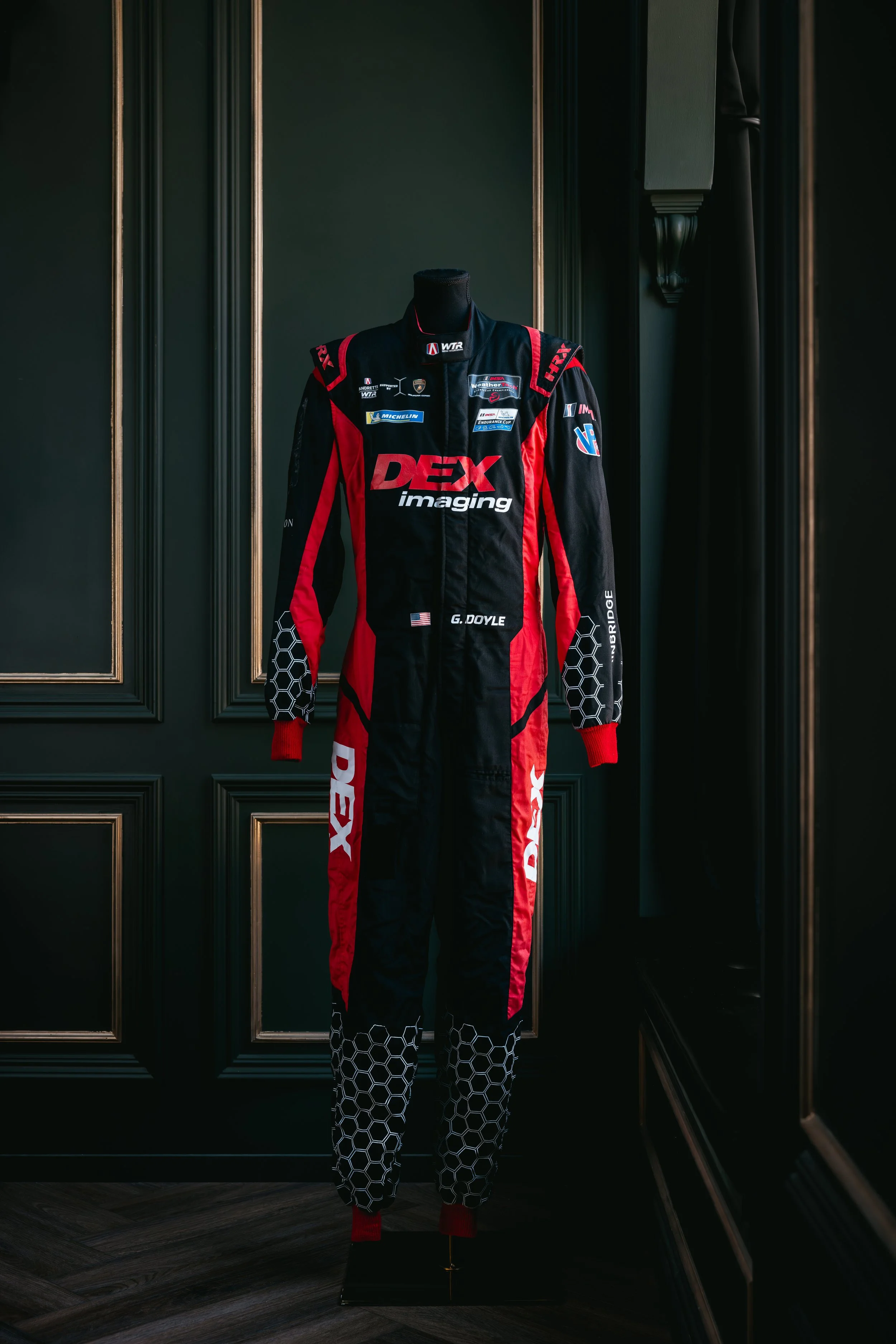 A race car driver standing on a brick-paved area with hands on hips, wearing a black and red racing suit, bright yellow racing shoes, and a white helmet with black and gold accents.
