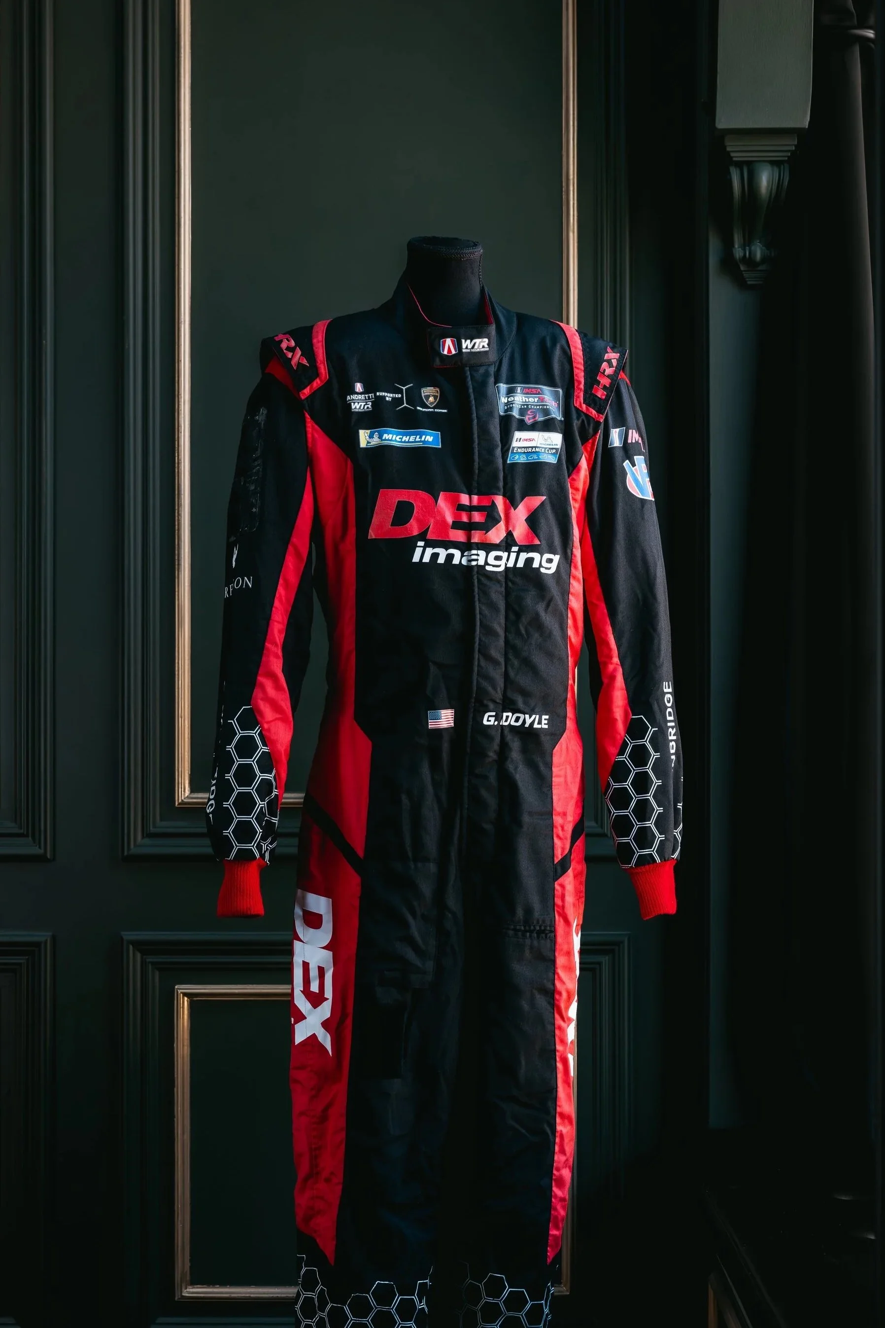 A race suit displayed on a black mannequin in front of dark green wall with gold accents.