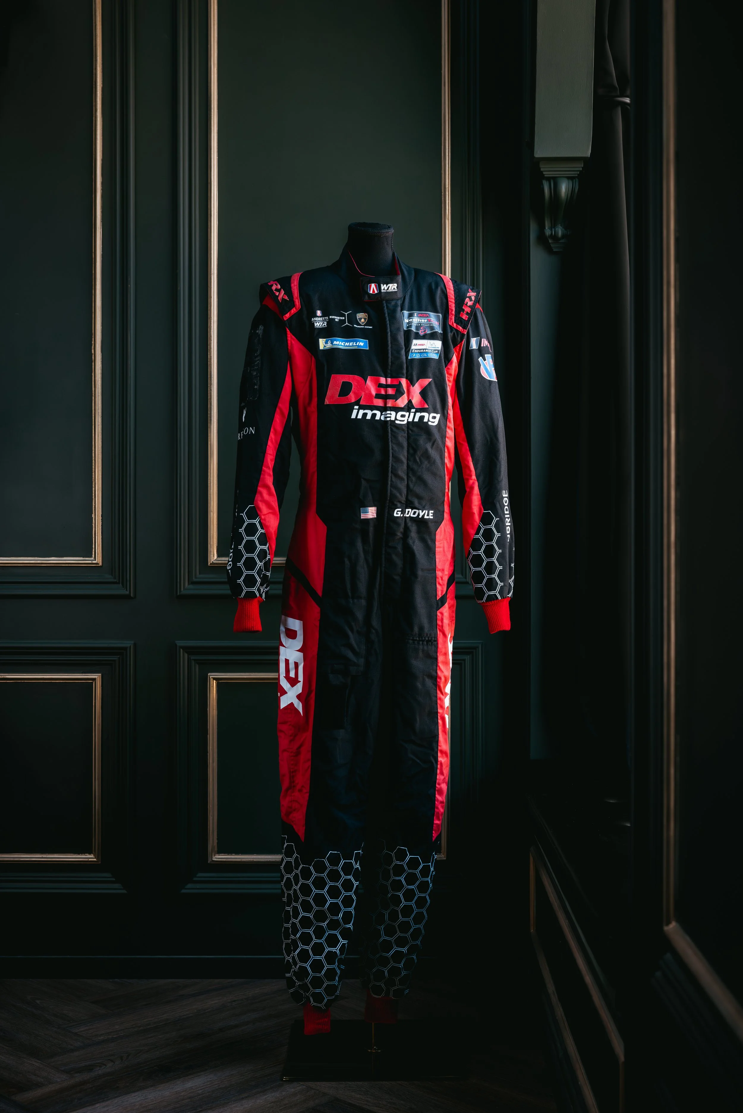 A racing suit with black, red, and white colors stands on a mannequin in front of a dark paneled wall. The suit features various sponsor logos, including DEX imaging, Michelin, and others.