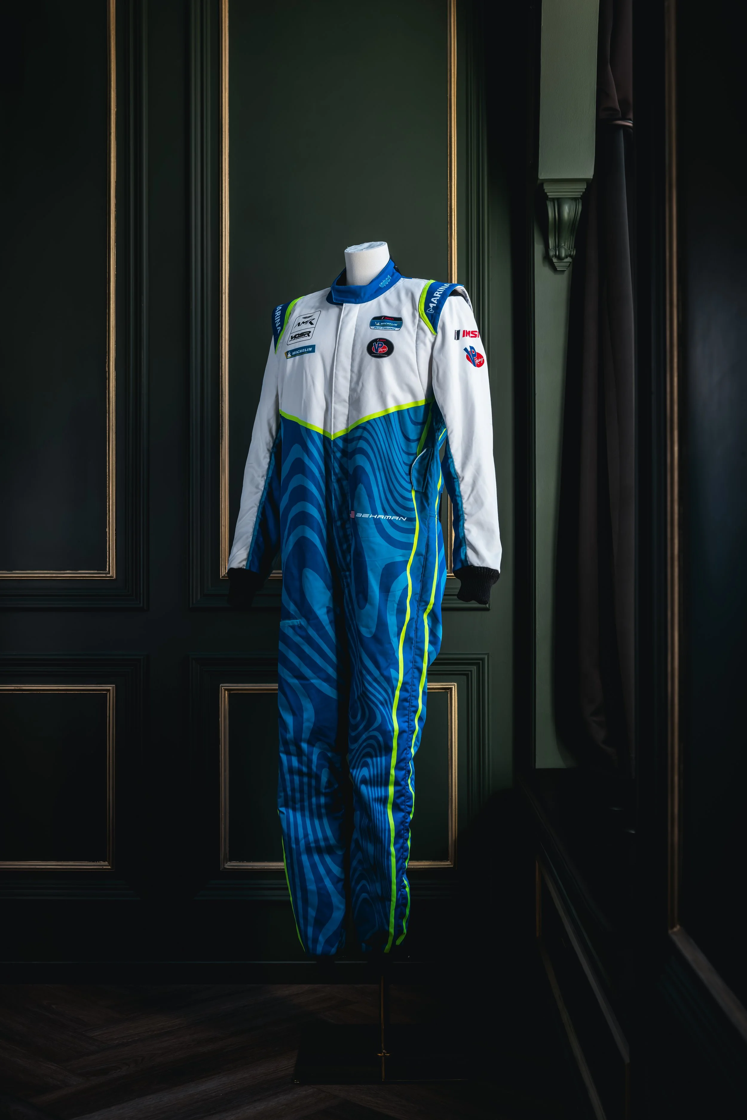 A racing suit on display, predominantly white and blue with neon yellow accents, displayed on a mannequin against dark green-paneled walls.