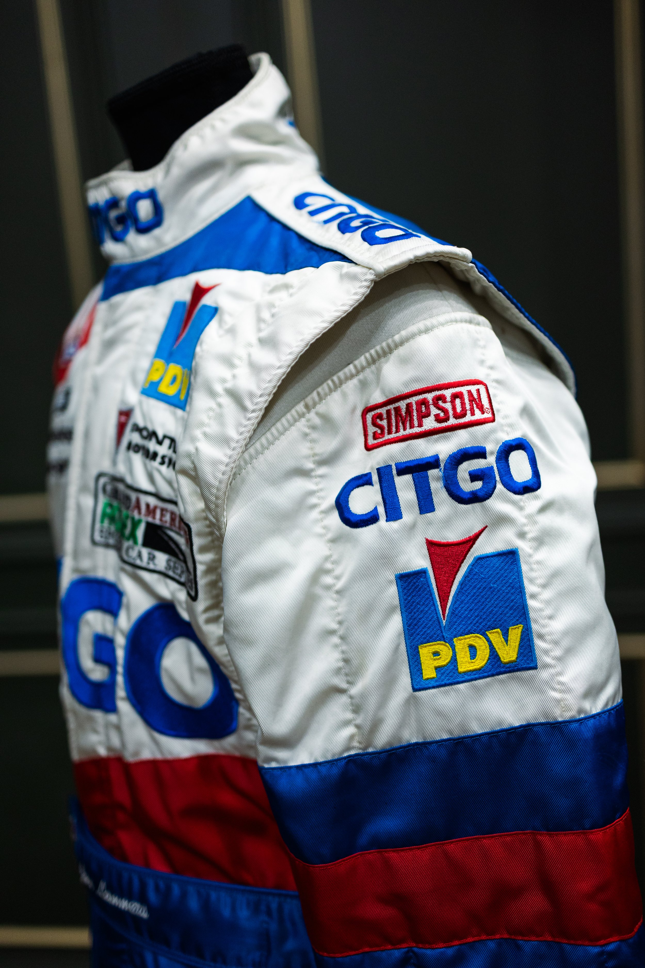 A white race car driver suit with multiple sponsor patches, including CITGO, PDV, and Simpson, displayed on a mannequin.