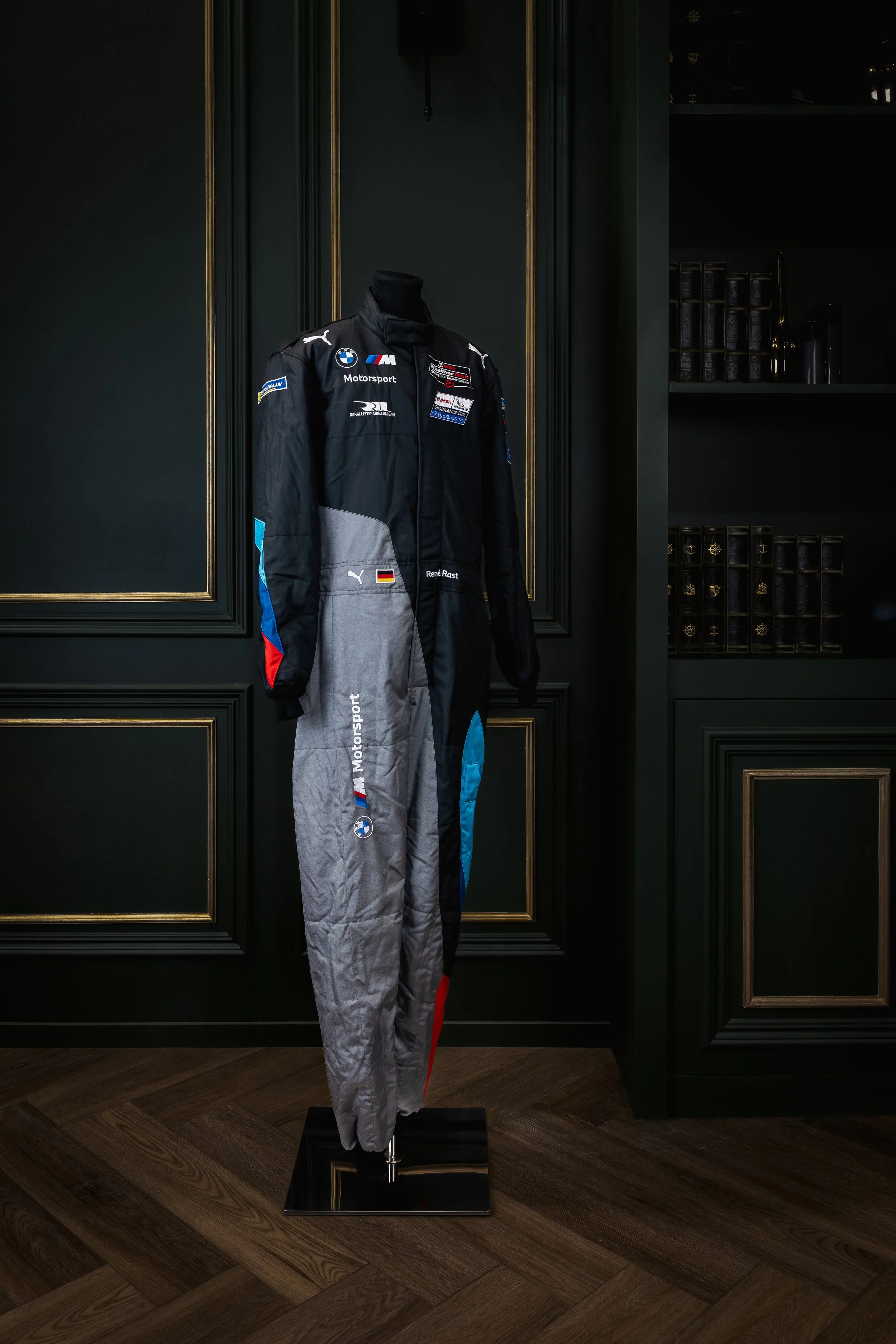 A racing suit on a mannequin display, featuring BMW Motorsport and PUMA logos, placed against a dark green wall with gold trim.