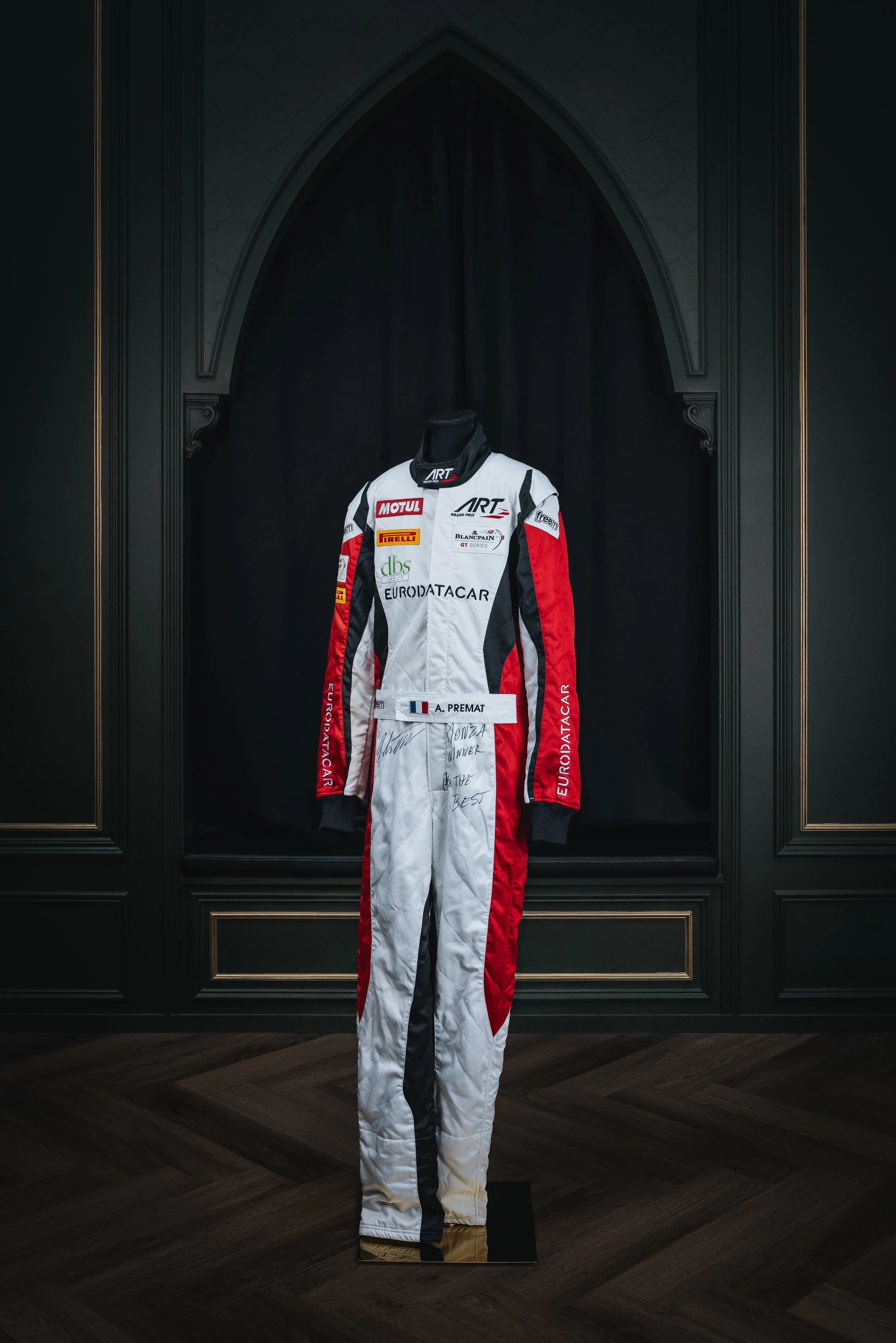 A racing suit on display in a glass case, signed and with a nameplate reading 'A. Premat.'