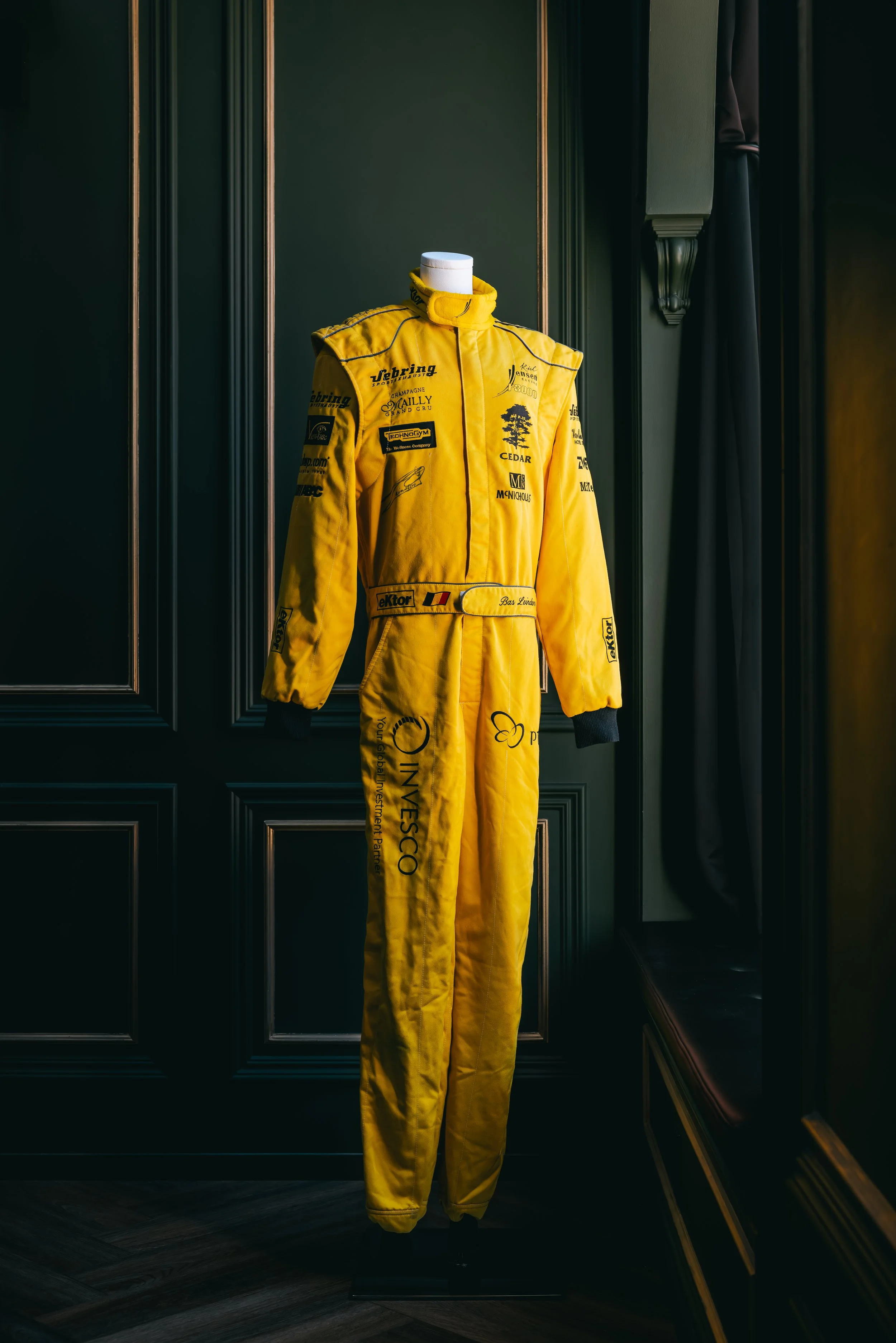 A yellow racing suit displayed on a mannequin against a dark wooden paneled wall.