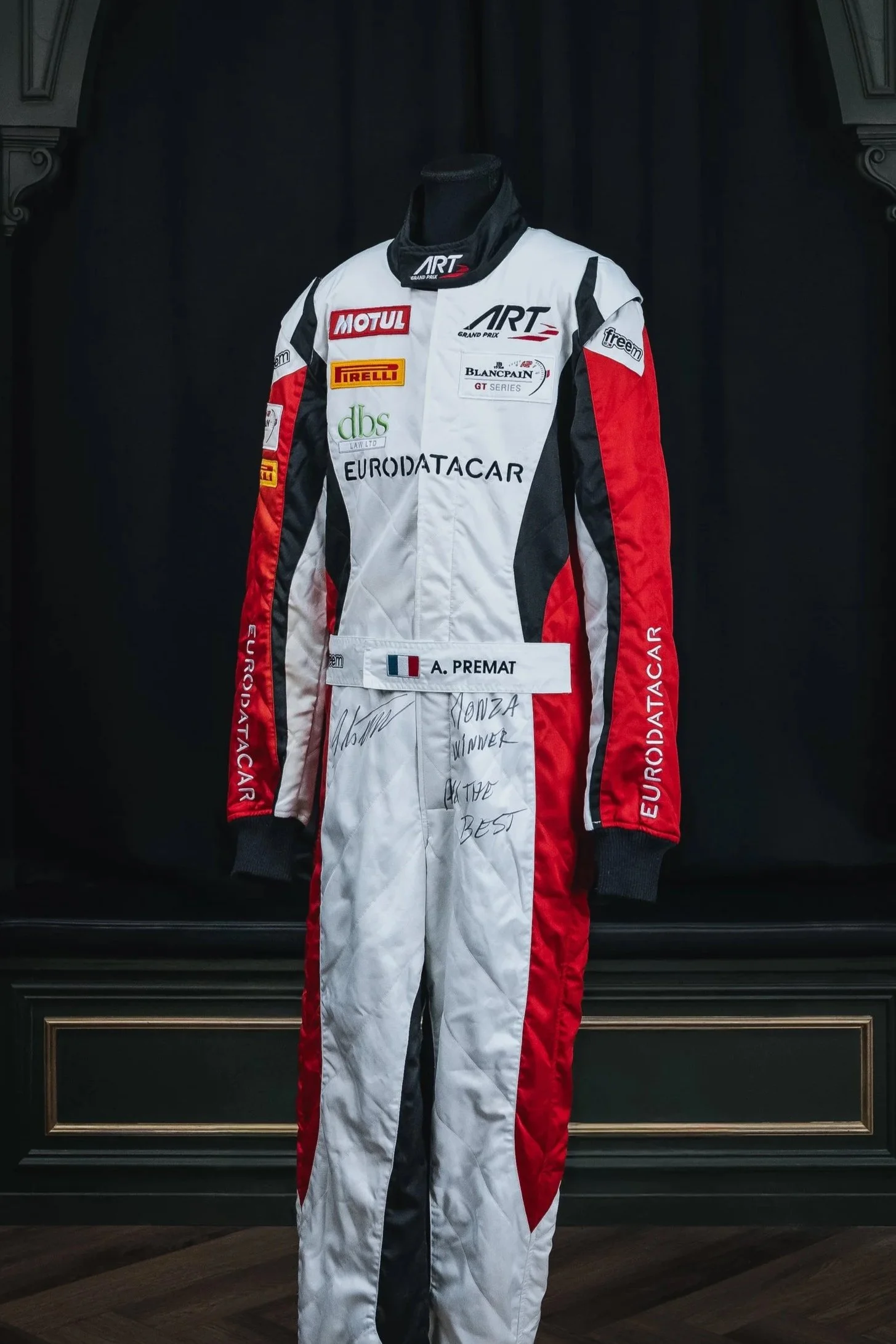 Racing suit on display, with various sponsor logos and a handwritten message, featuring a French flag and the name A. Premat.