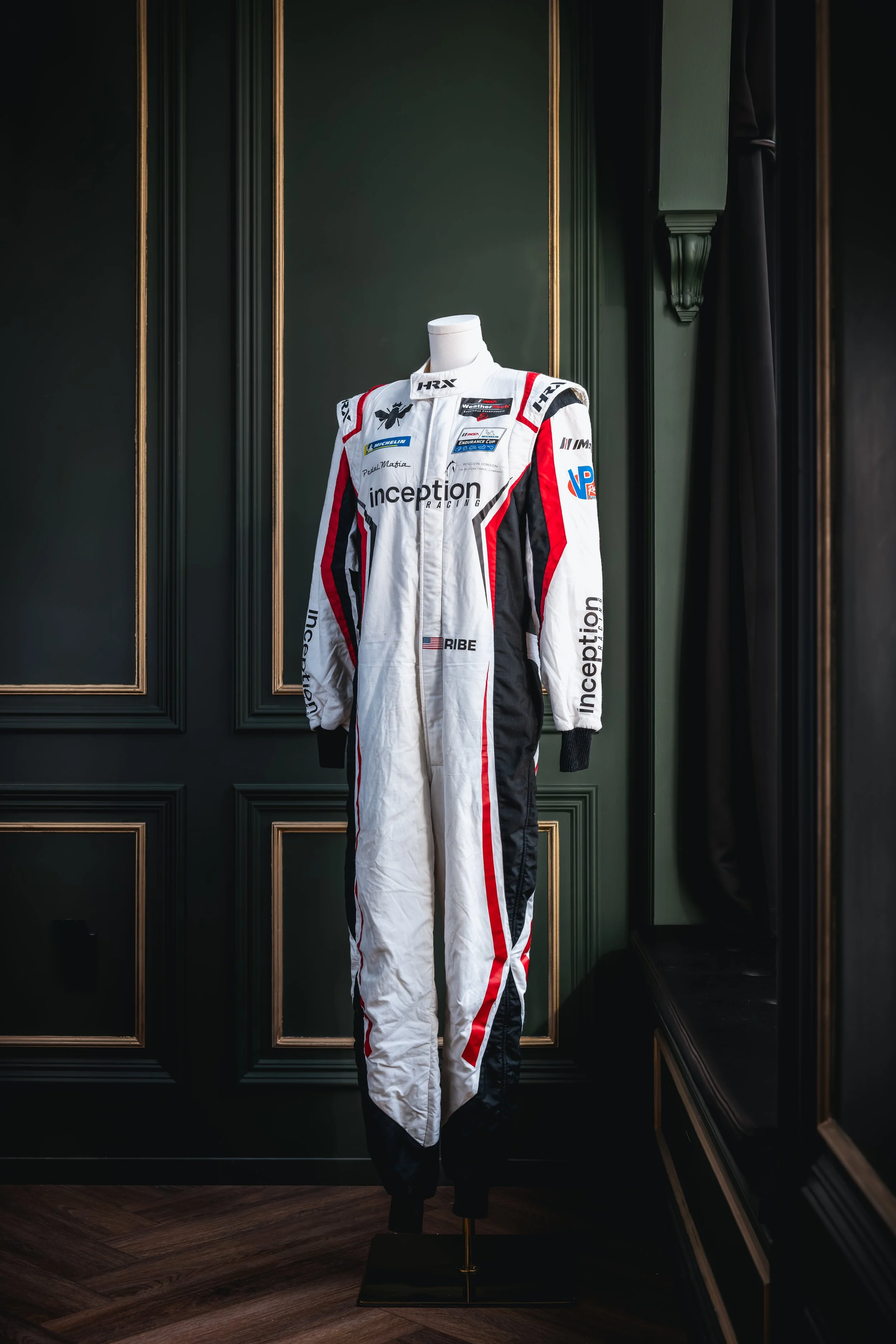 A racing driver’s suit on a mannequin, featuring various sponsor patches, predominantly in white, black, and red, displayed against a dark green paneled wall.
