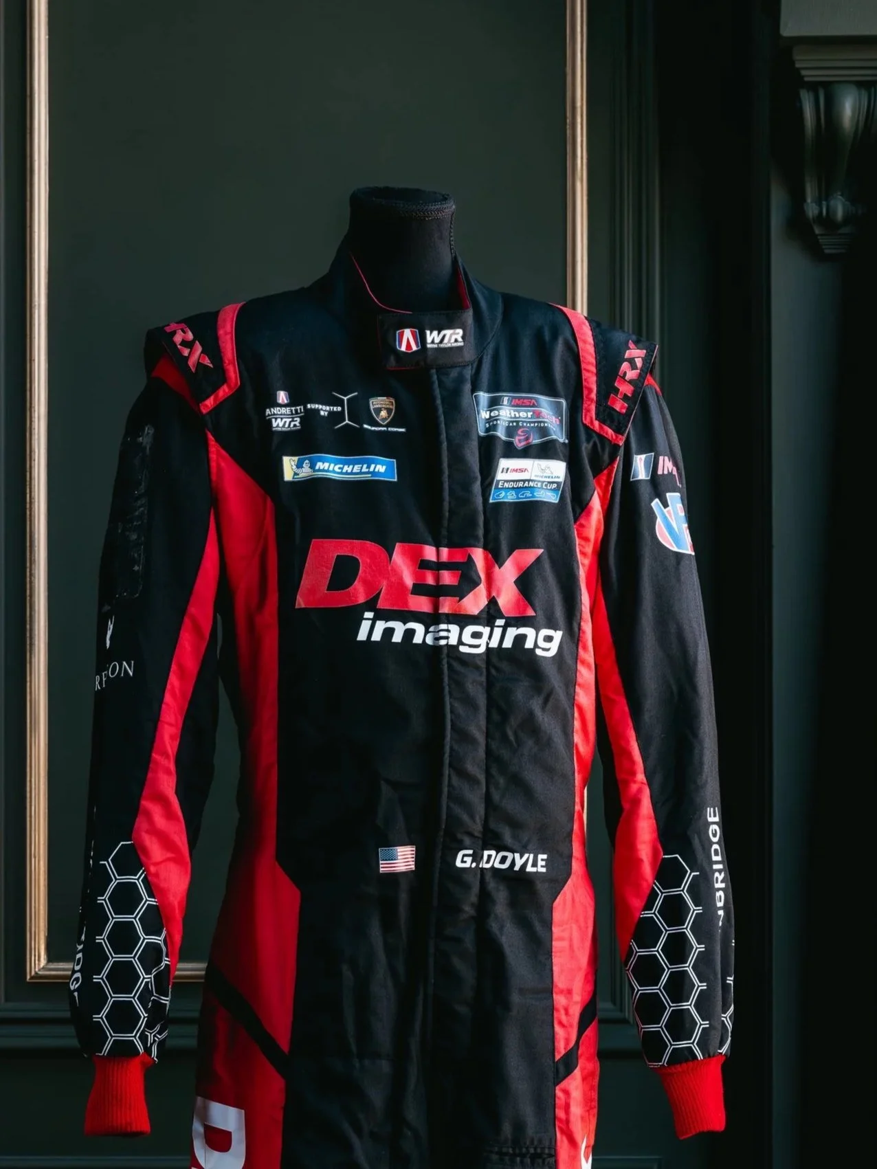 A racing suit displayed on a mannequin, black and red with various sponsor logos, including DEX imaging and Michelin.