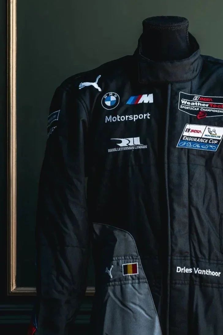 Black racing jacket with sponsor patches, including BMW, Puma, and Michelin, displayed on a mannequin. The jacket features a Belgian flag patch on the sleeve and the name 'Dries Vanthoor' embroidered on the chest.