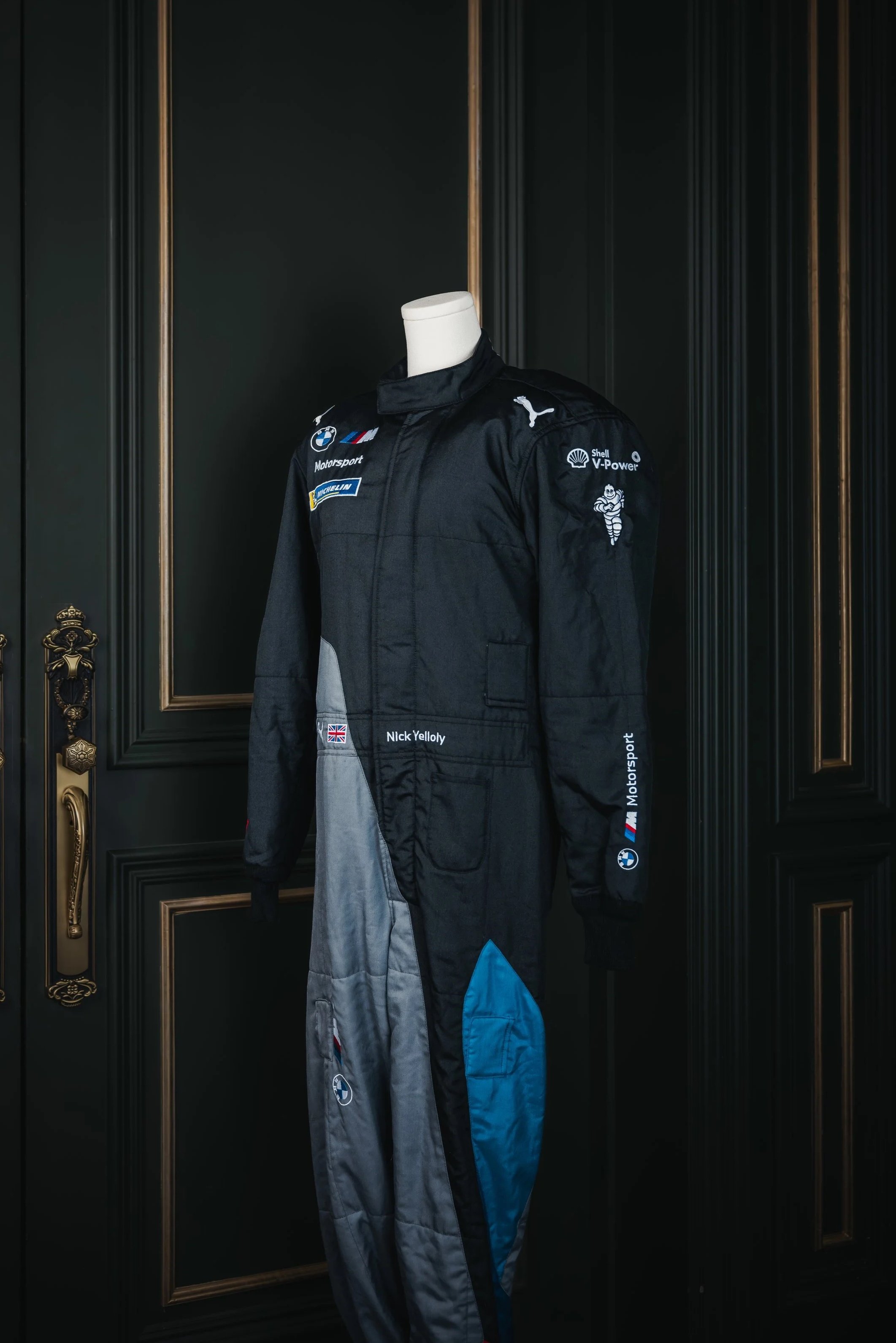 A black racing suit on a mannequin, with sponsor patches including BMW, Puma, Michelin, Shell, and Michelin, displayed against a dark paneled wall.