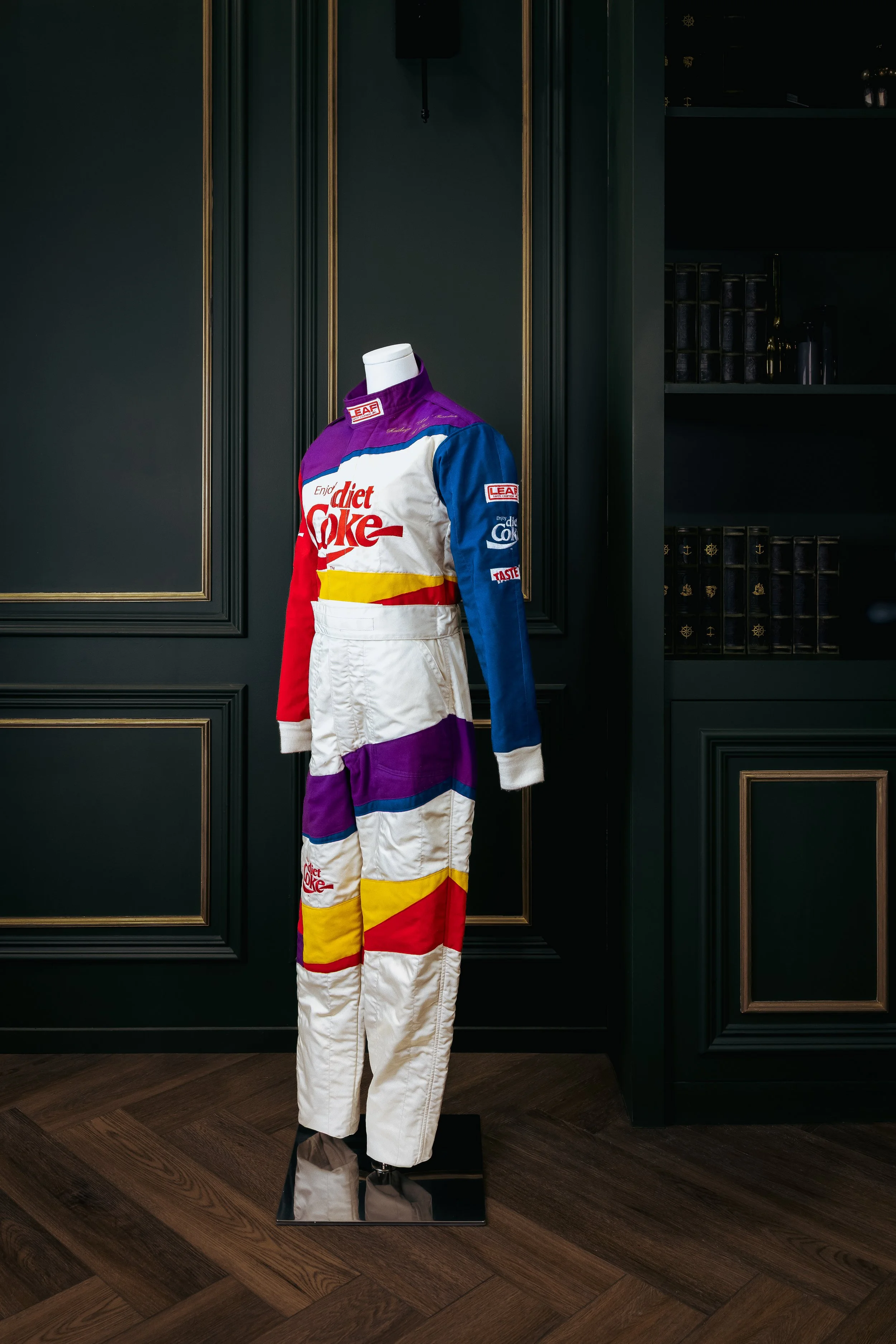 Colorful racing suit with logos, displayed on a mannequin in a room with dark green walls and wood floors.