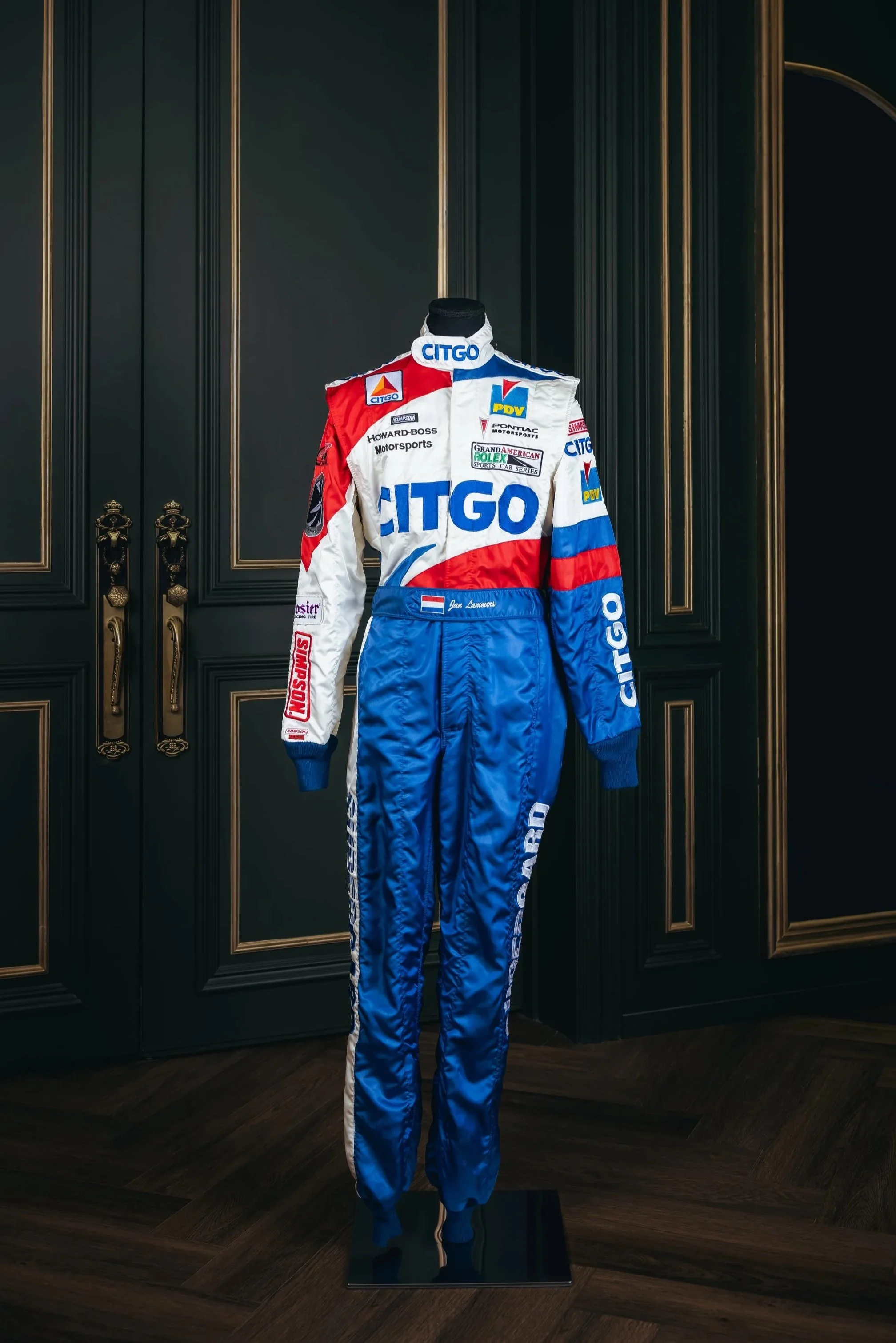 A vintage racing suit displayed on a mannequin in a luxurious room with dark wood paneling. The suit is primarily white with red and blue accents, featuring sponsor logos including Citgo and others.
