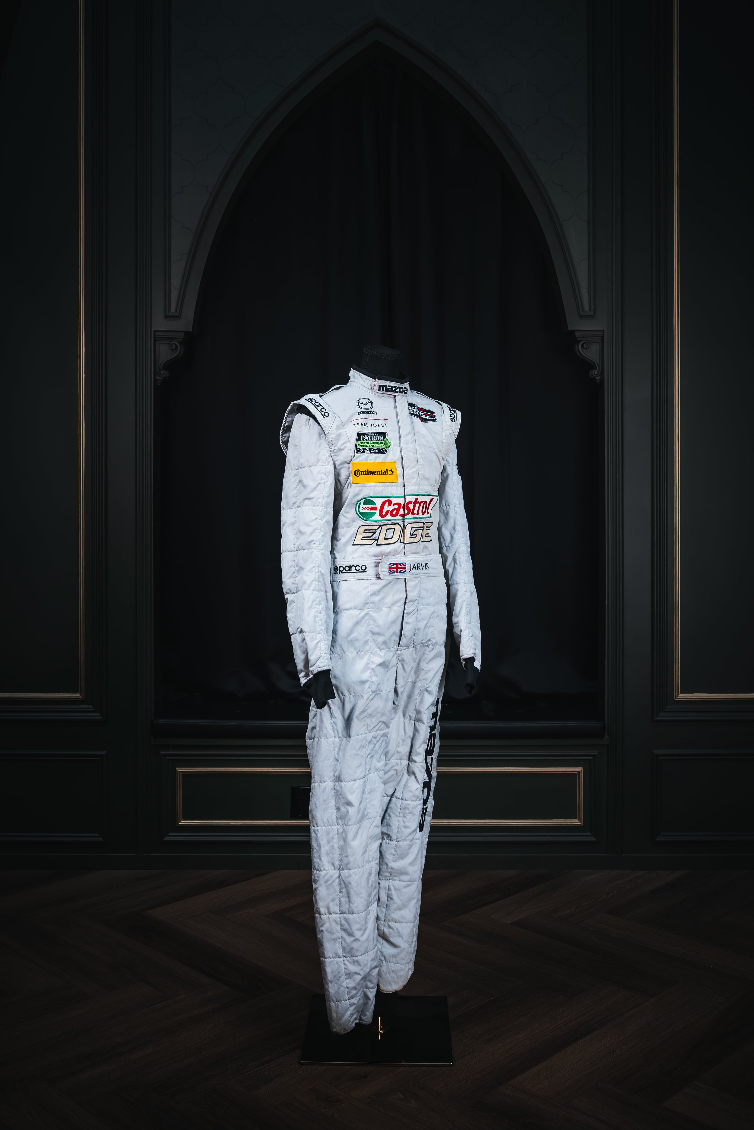 A white racing suit with various sponsor logos displayed on a black mannequin in a dark room with decorative black walls.