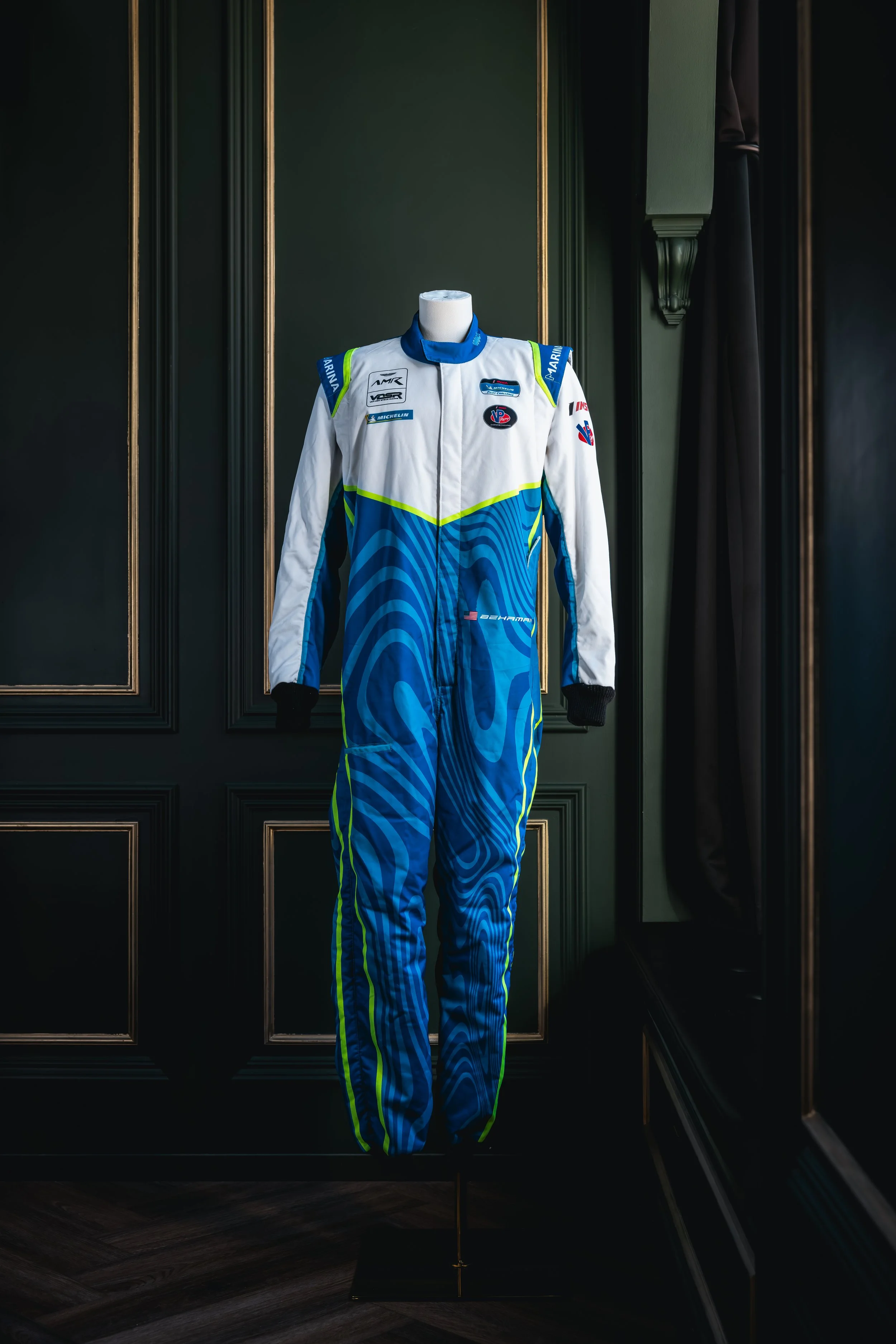 A racing driver’s suit displayed on a mannequin against a dark green paneled wall.