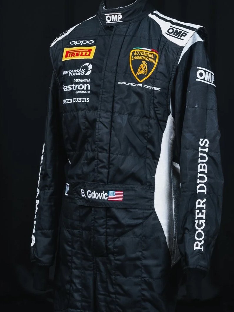A racing suit with Lamborghini and sponsor logos, including Pirelli, Pertamina, and Astron, on a black background. The suit has a Lamborghini badge and the name B. Govic on the belt with an American flag.