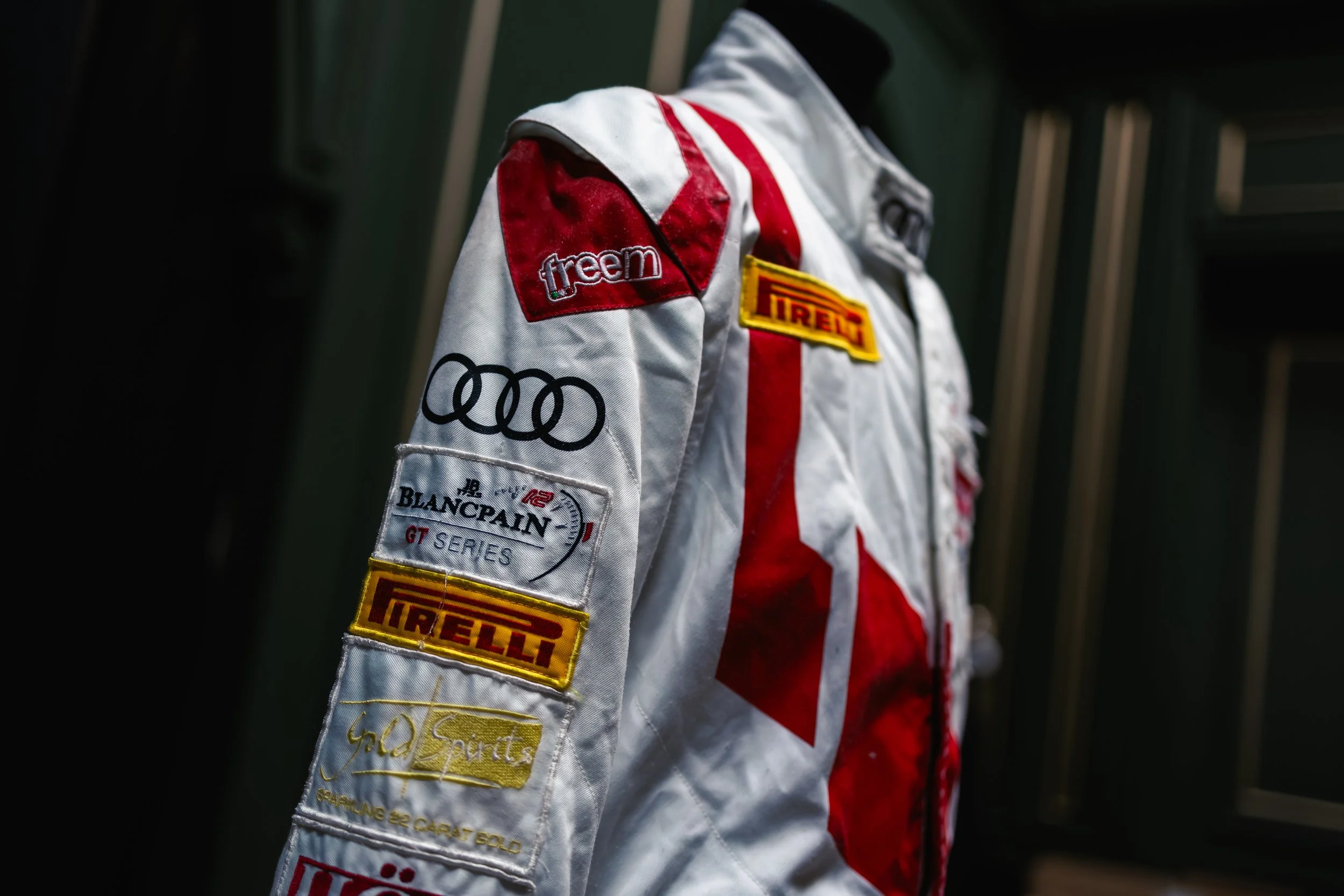A racing suit with sponsor patches, including Pirelli, Audi, Blancpain, and others, displayed inside a dark room or locker.