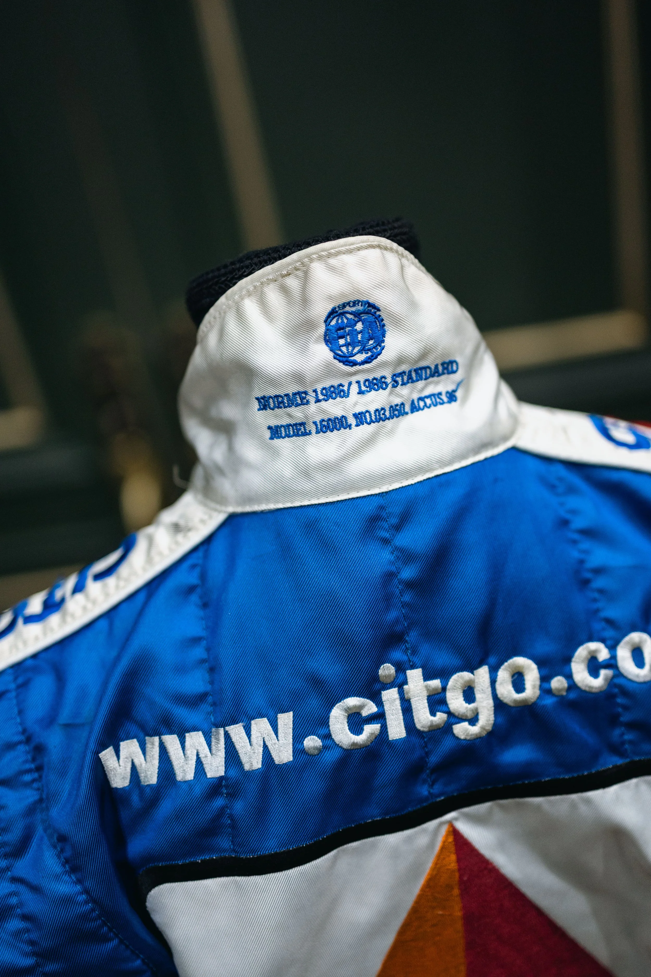 The photo shows a racing jacket with the website 'www.citgo.com' and a white collar with blue embroidery that mentions 'WORLD RACING 1986/1986 STANDARD MODEL 1600L, MOTOR, ACTUS SP.' The jacket features the CITGO logo and a multicolored geometric design.