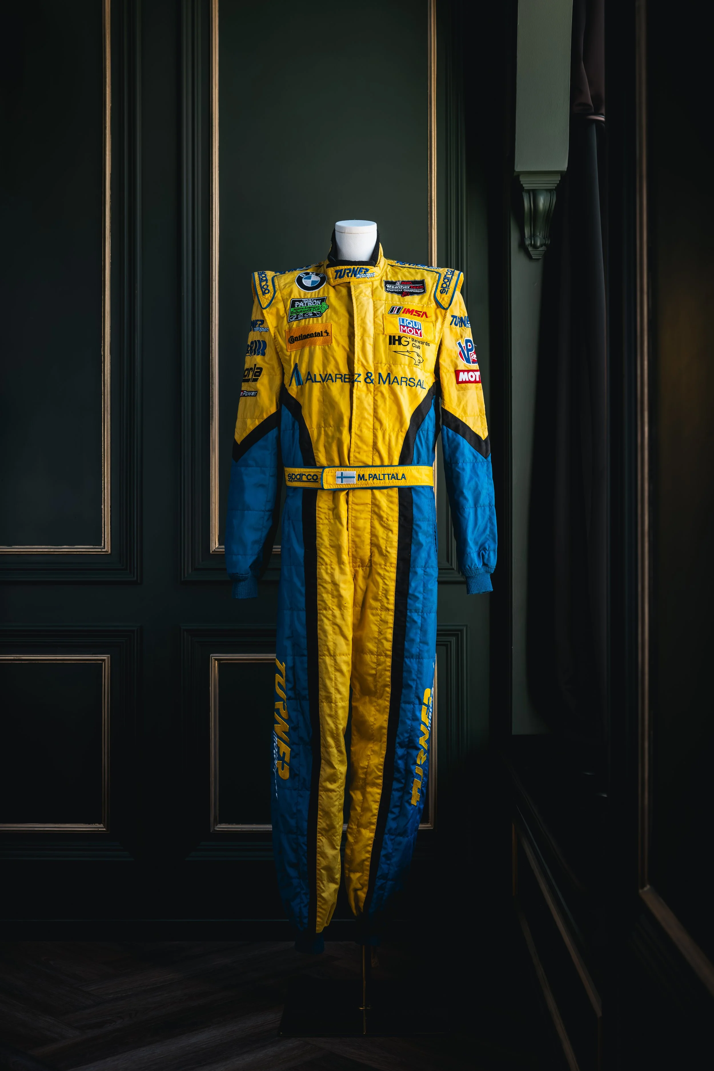 A yellow and blue racing suit with various sponsor patches displayed on a mannequin in a dark room with paneled walls.