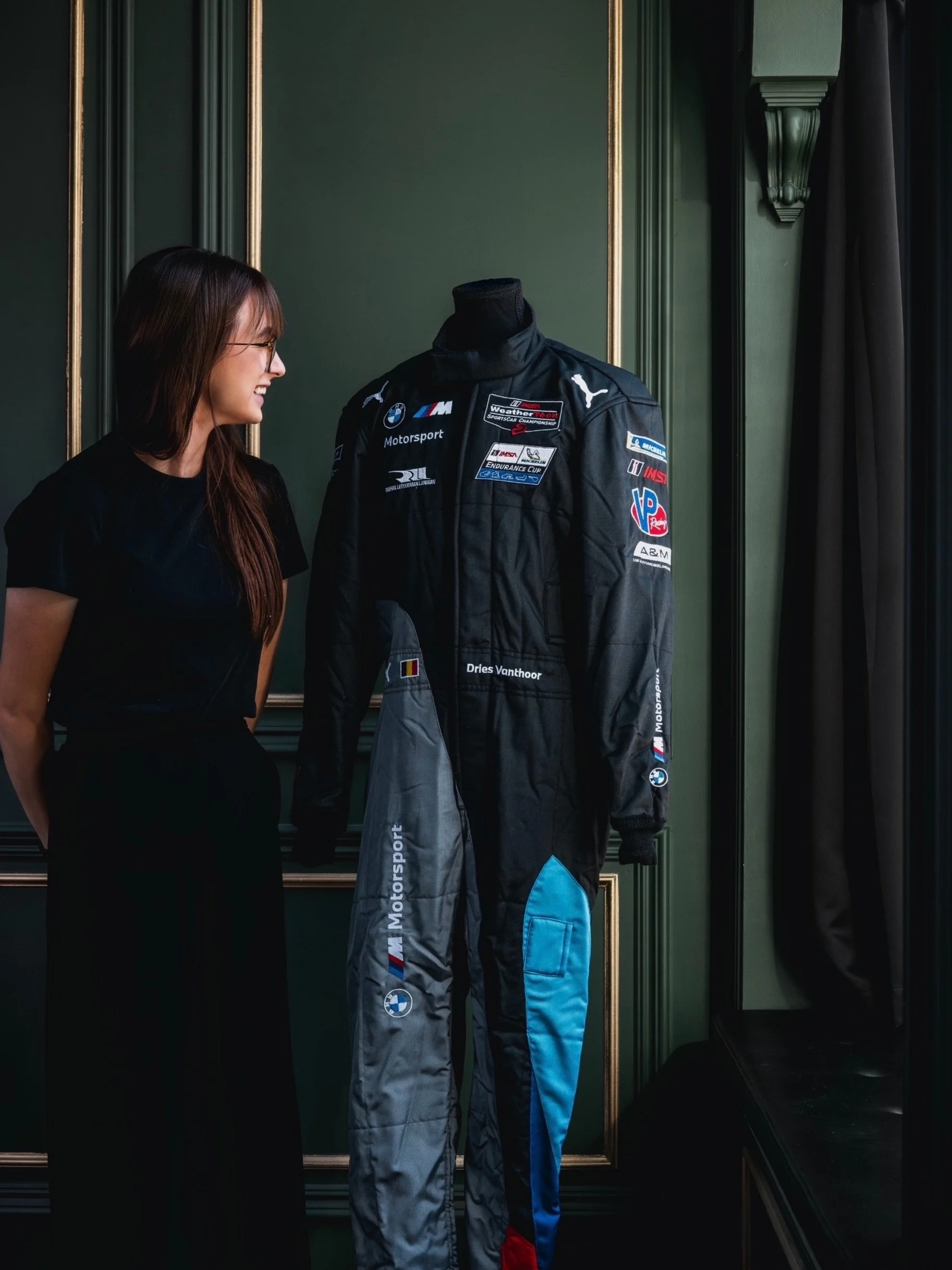 A woman with glasses and long brown hair looking at a racing suit on a mannequin. The suit is black with various sponsor patches and the name 'Dries Vanthoor'. The woman is smiling and standing in a room with dark green walls and gold trim.