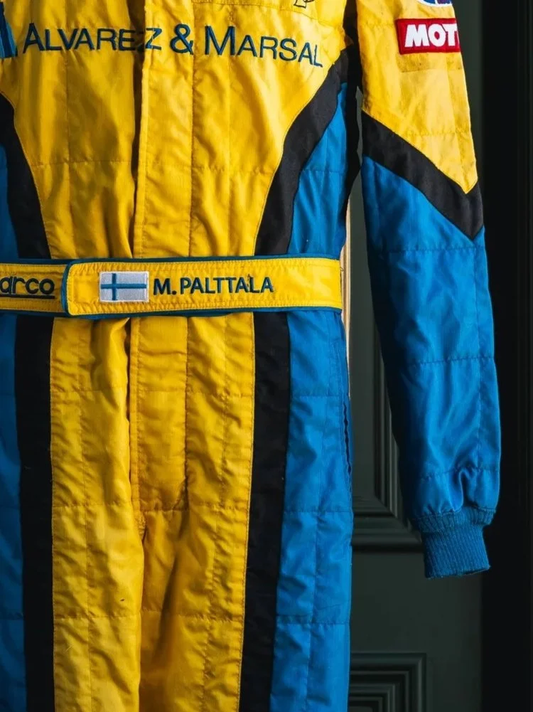 A racing driver suit in yellow and blue with patches and a name tag reading 'M. Palttala', featuring a Finnish flag, and various sponsor patches.