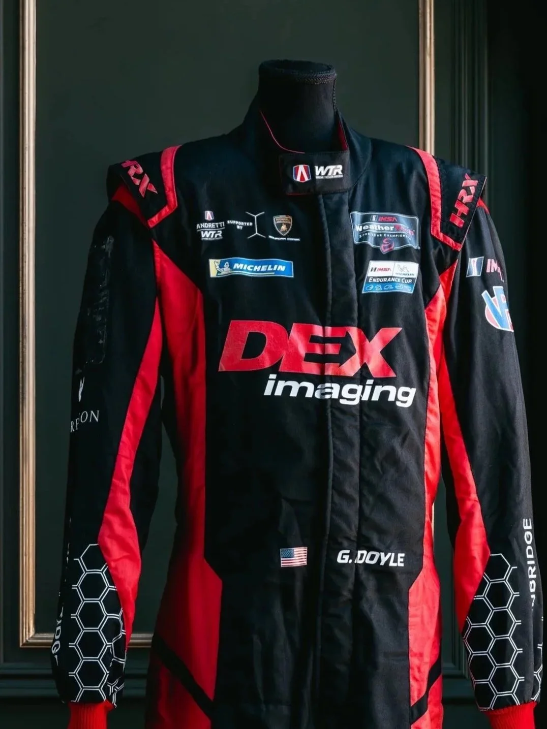Race car driver's racing suit with sponsor logos, including DEX imaging, Michelin, IMSA, and American flags, displayed inside a frame.