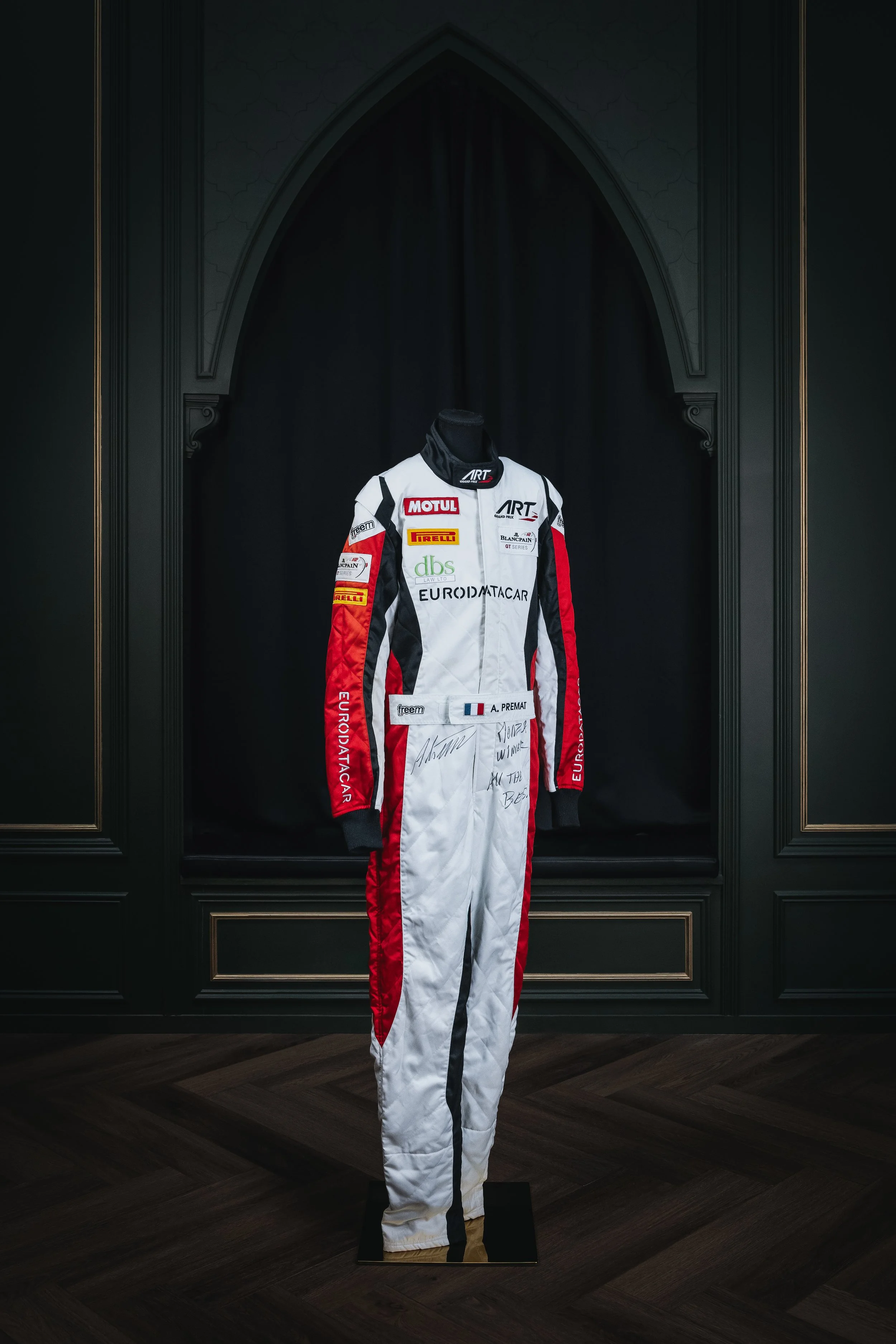A racing suit displayed on a mannequin against a black backdrop in an ornate room with dark wood flooring.